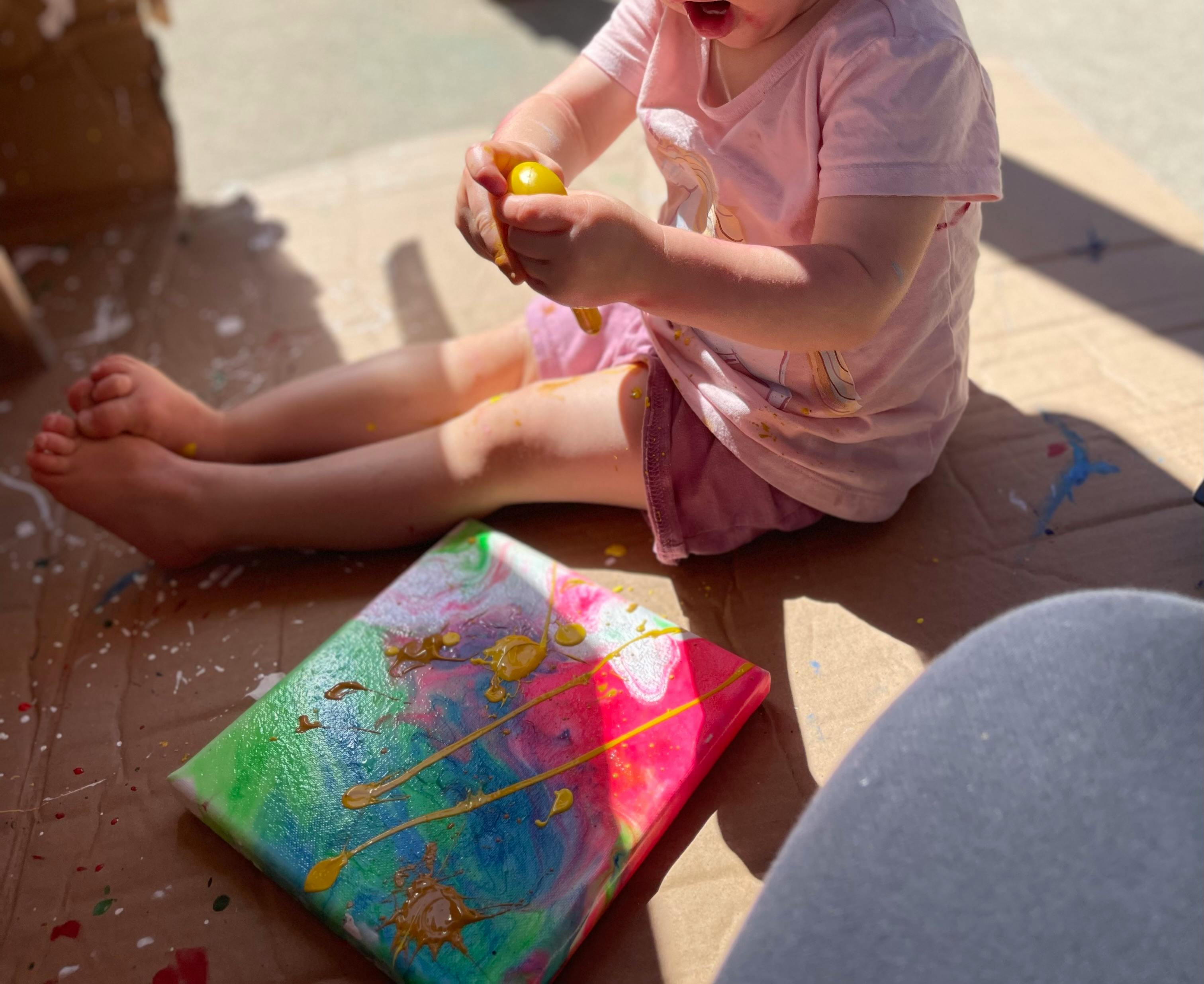 Child sitting on the ground holding a yellow object, next to a colorful painting