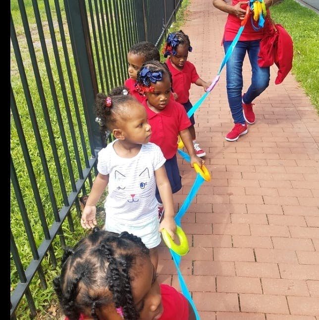 Children holding onto a blue rope with handles, walking on a brick path
