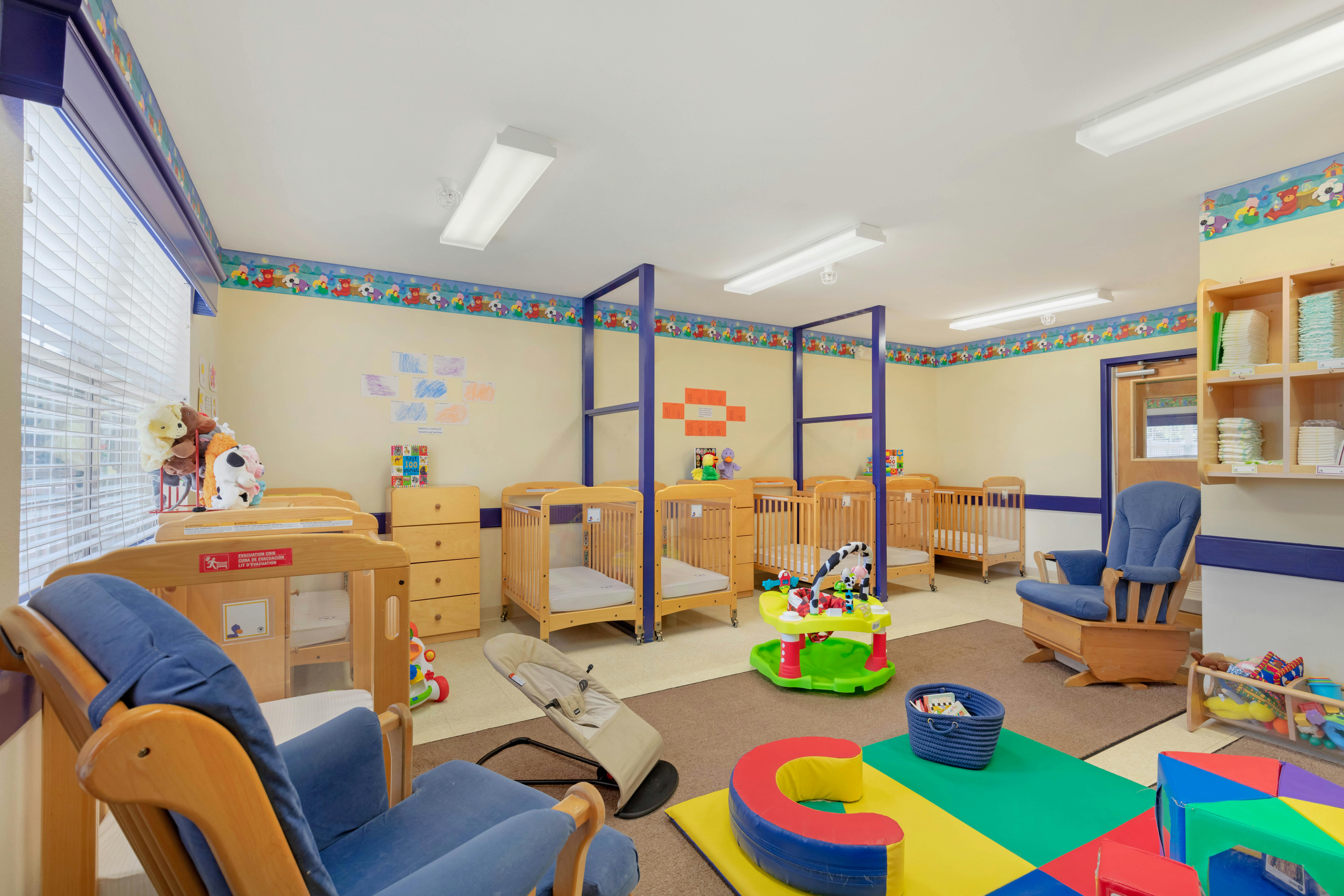 A colorful childcare room with cribs, chairs, and baby toys