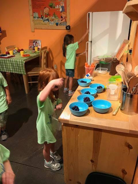 Child reaching for a refrigerator, another child standing near bowls of blueberries