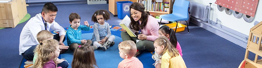 Two adults reading to a group of children sitting on the floor in a classroom