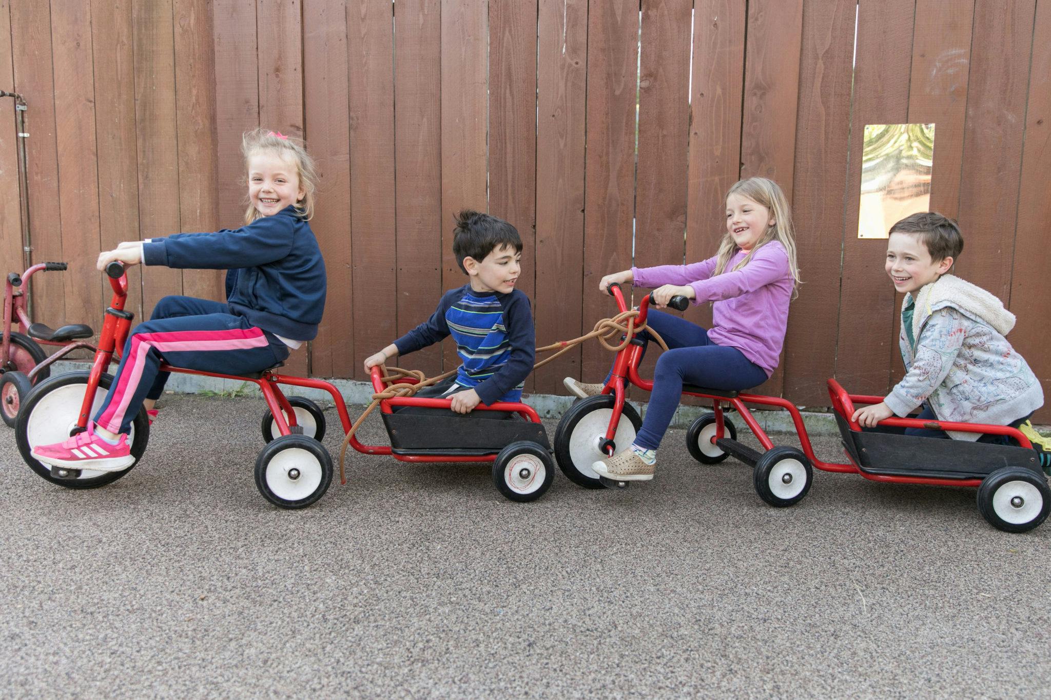 Four children riding connected tricycles with wagons
