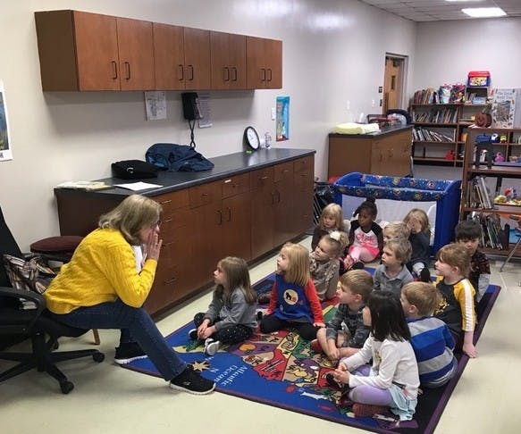 Person speaking to a group of children sitting on a rug in a classroom