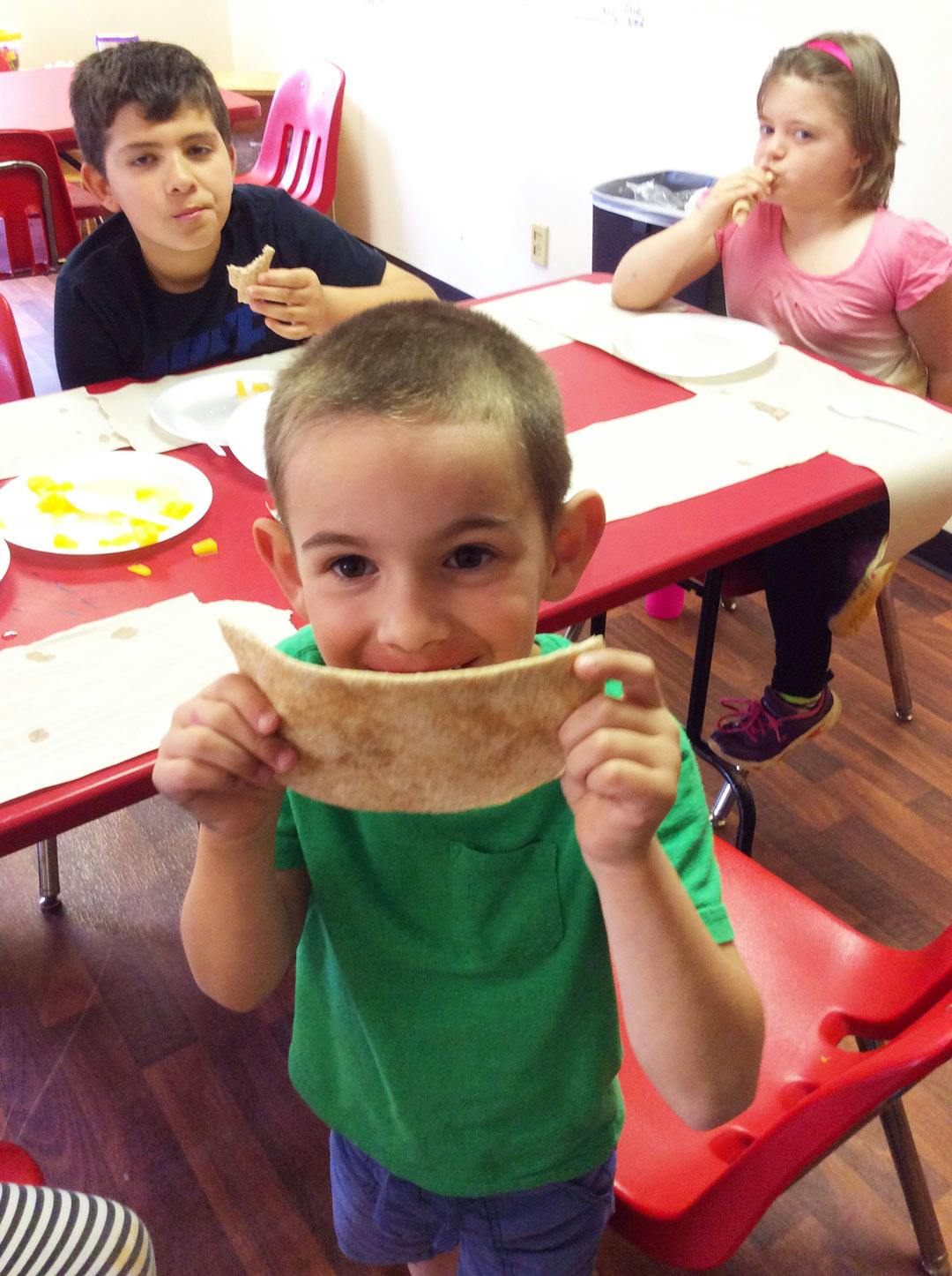 Child holding a flatbread in front of their face, with other children at a table