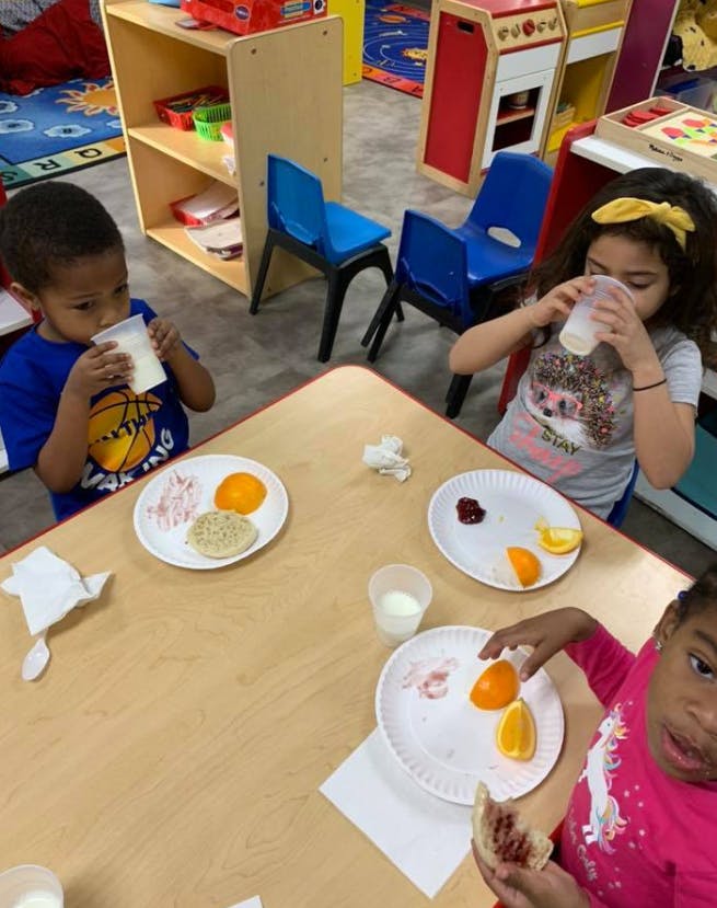 Three children eating and drinking at a table in a classroom