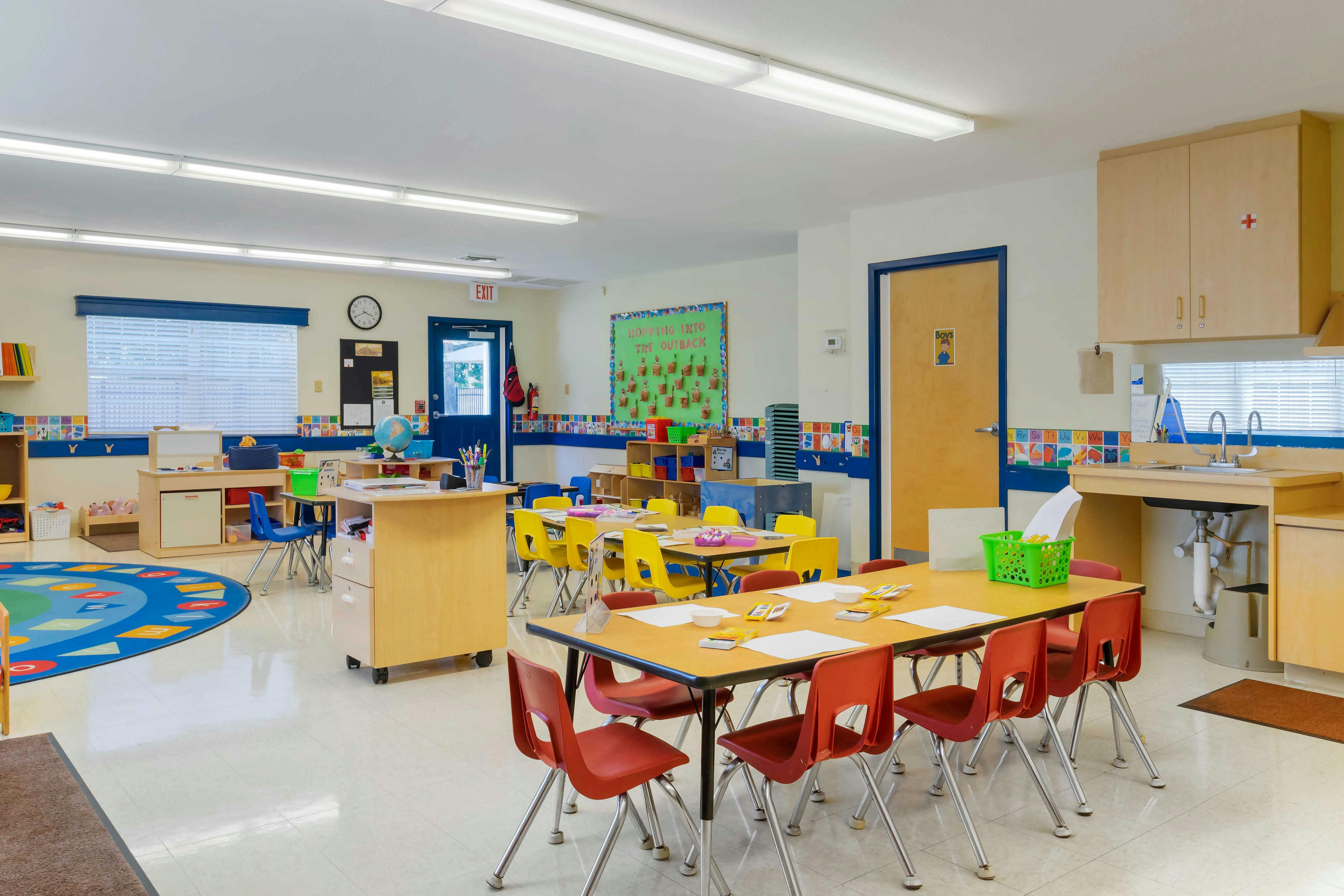 An empty classroom with several tables and chairs, a sink, and an exit door