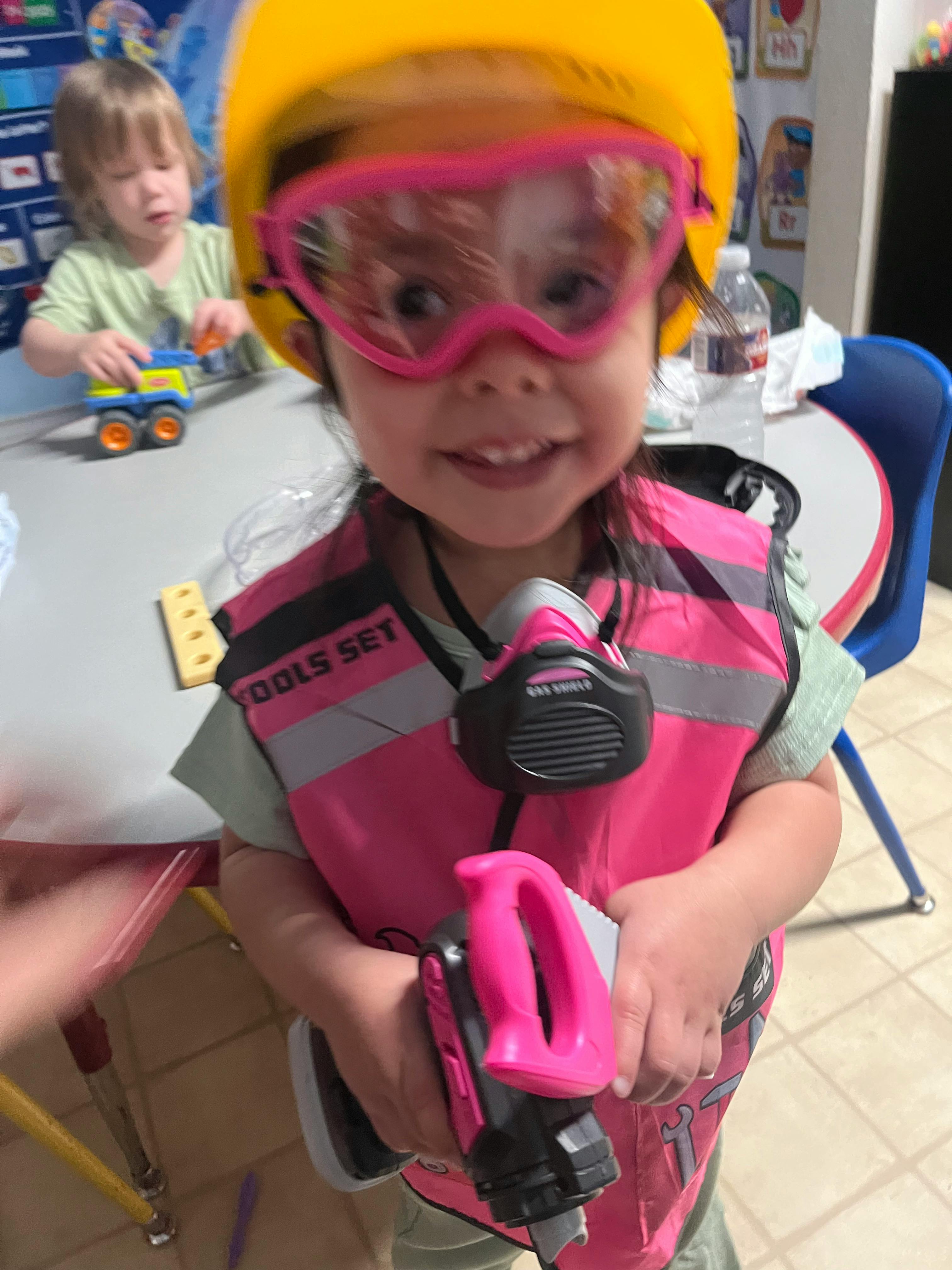 Child wearing a yellow hard hat, pink safety goggles, and a pink vest, reflective vest