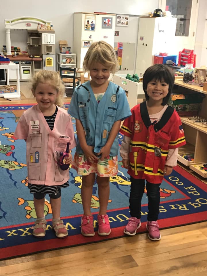 Three children in dress-up costumes standing on a rug in a classroom