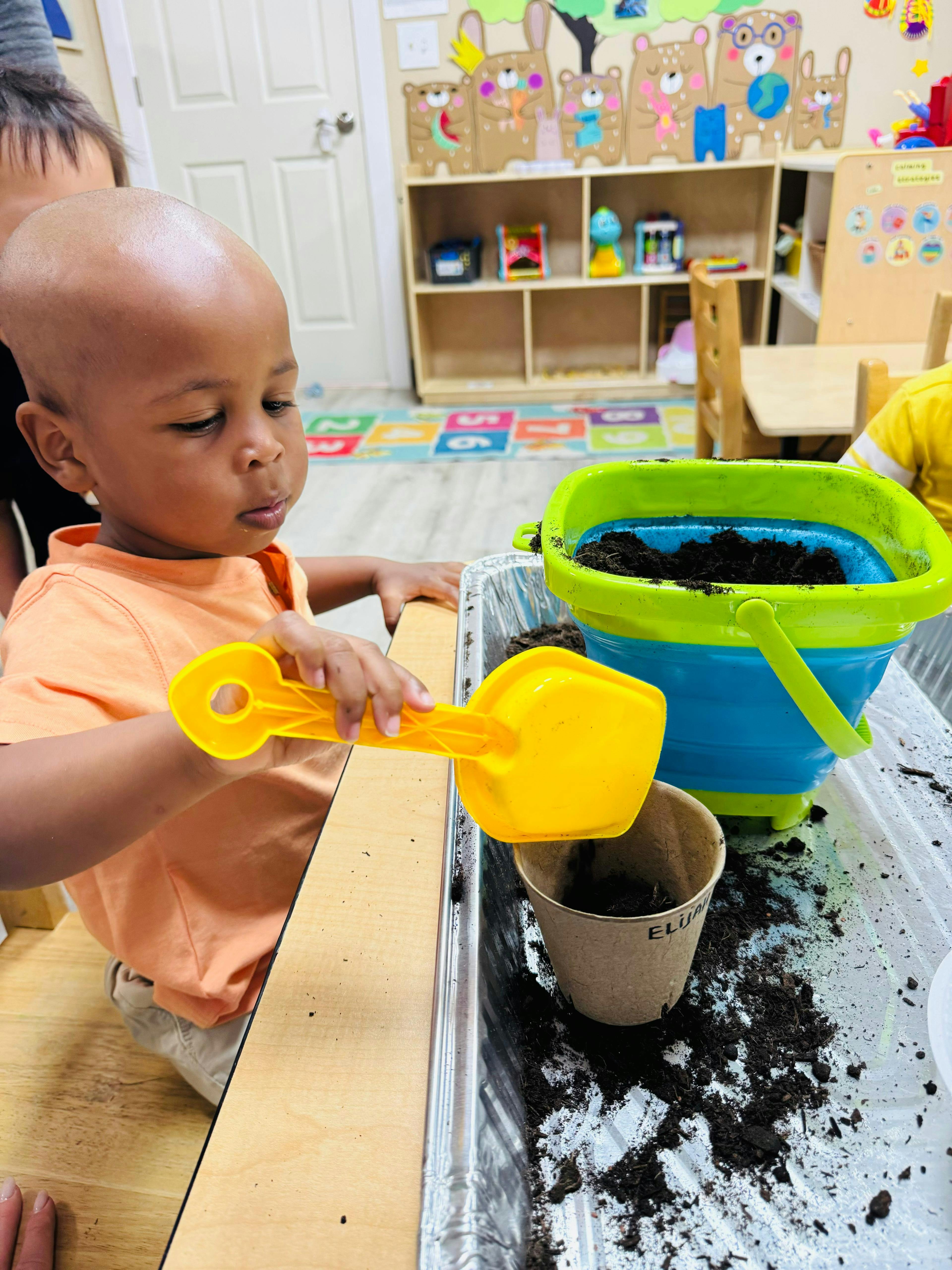 Child using a yellow scoop to fill a paper cup with soil in a classroom