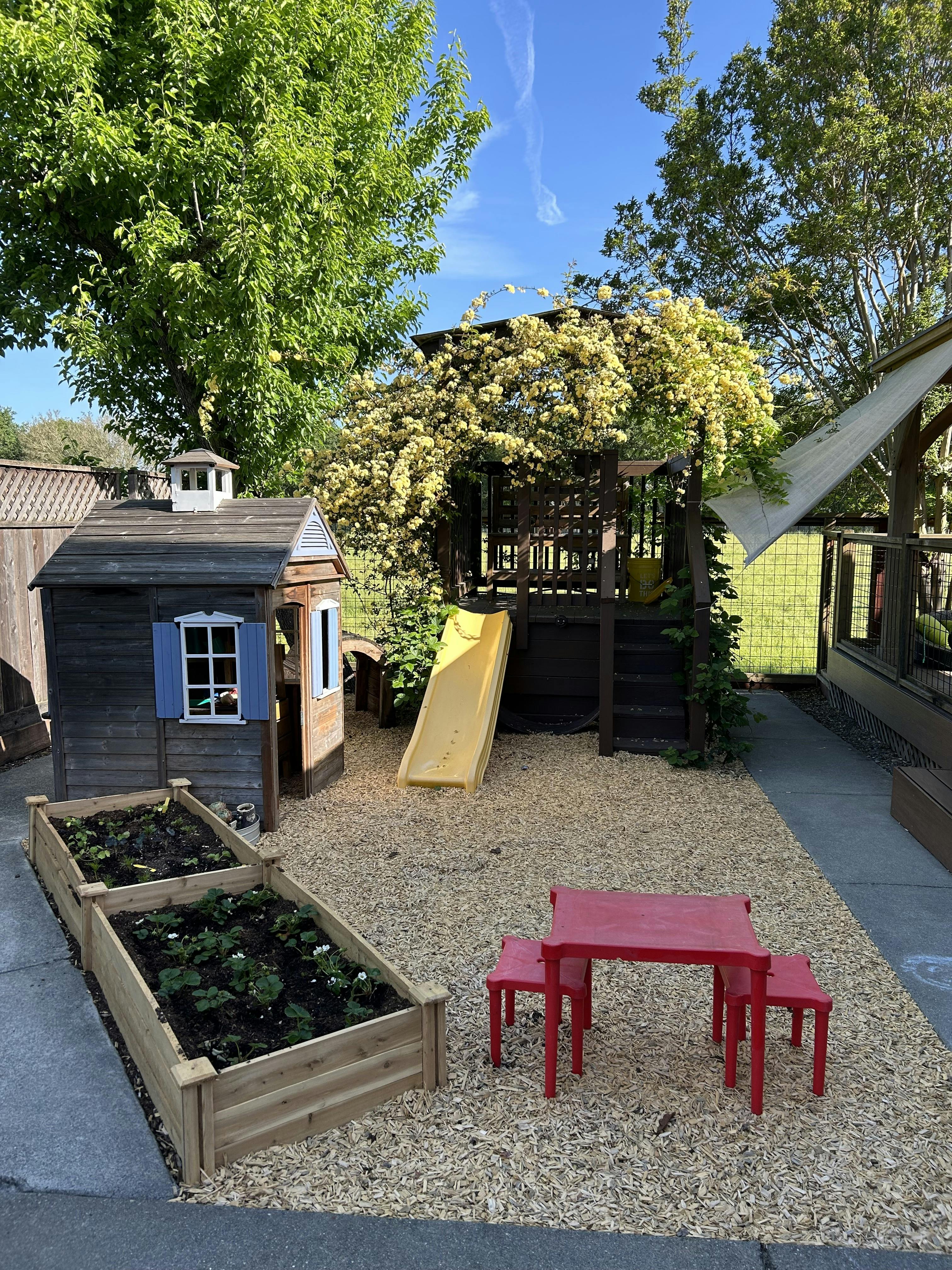 Playground with a wooden playhouse, slide, raised garden beds, and red table