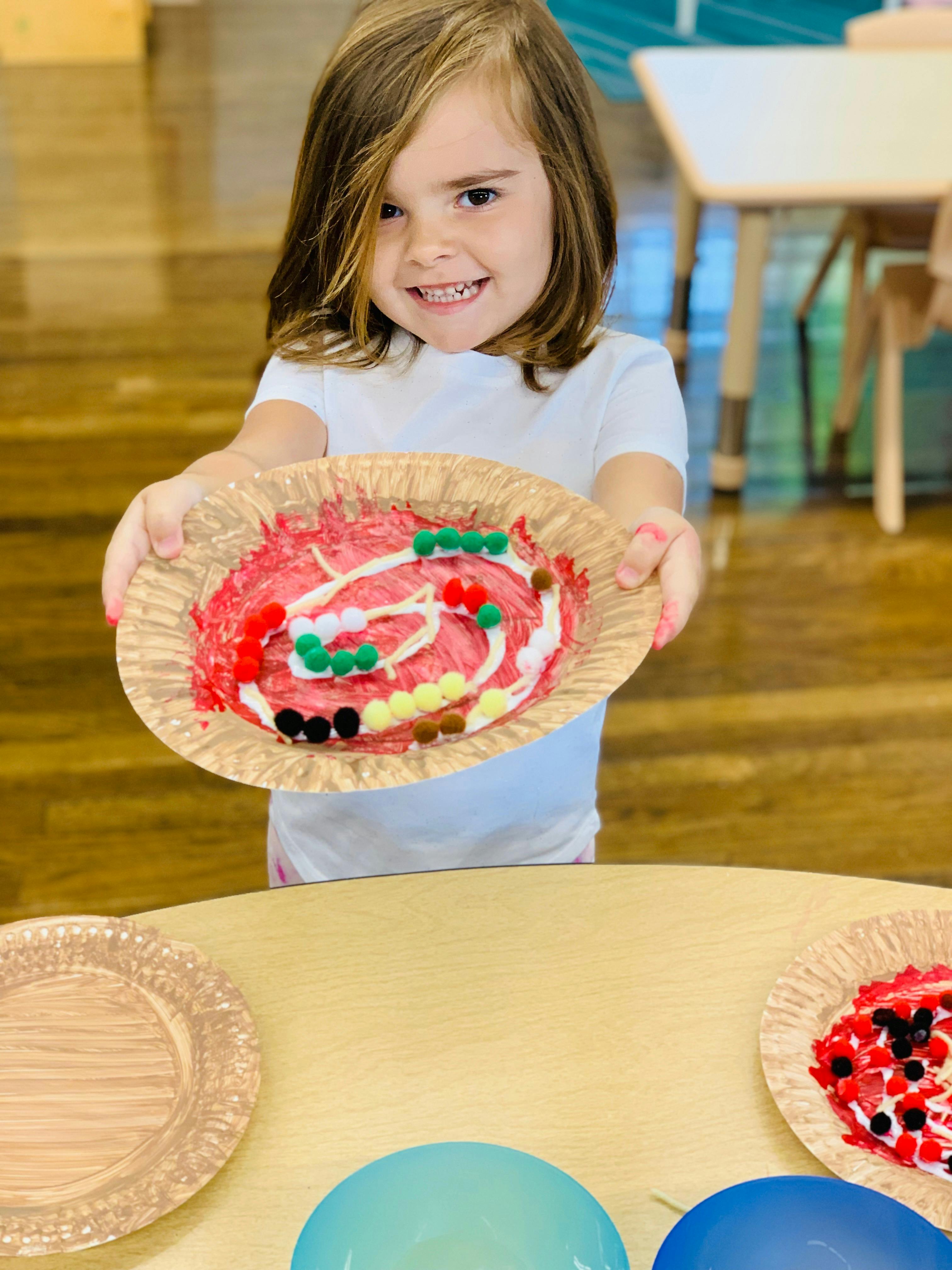Child holding up a paper plate with a craft project made of pom-poms and paint