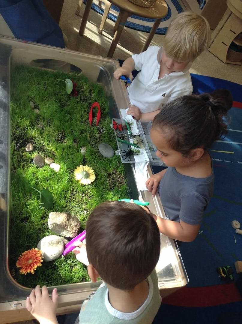 Three children playing with a sensory bin filled with grass and toys
