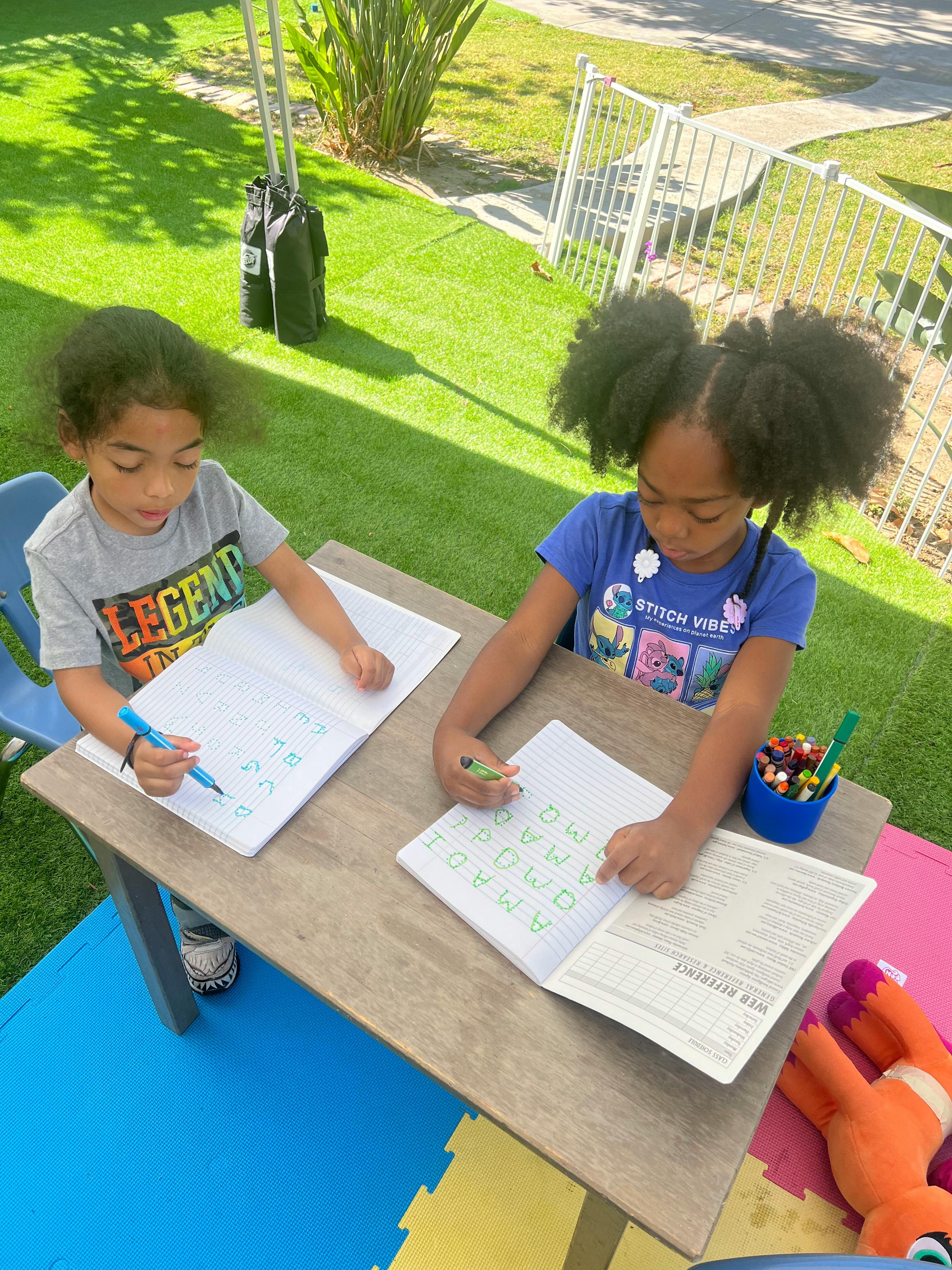 Two children writing in notebooks at a table outdoors