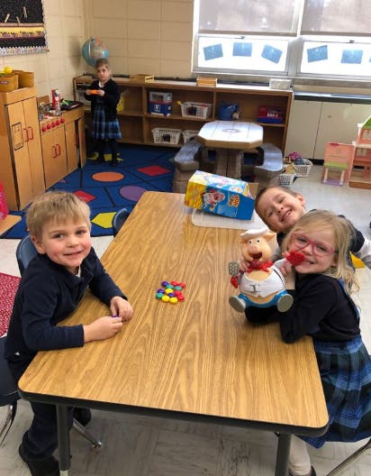 Three children in a classroom, two at a table with colorful counters