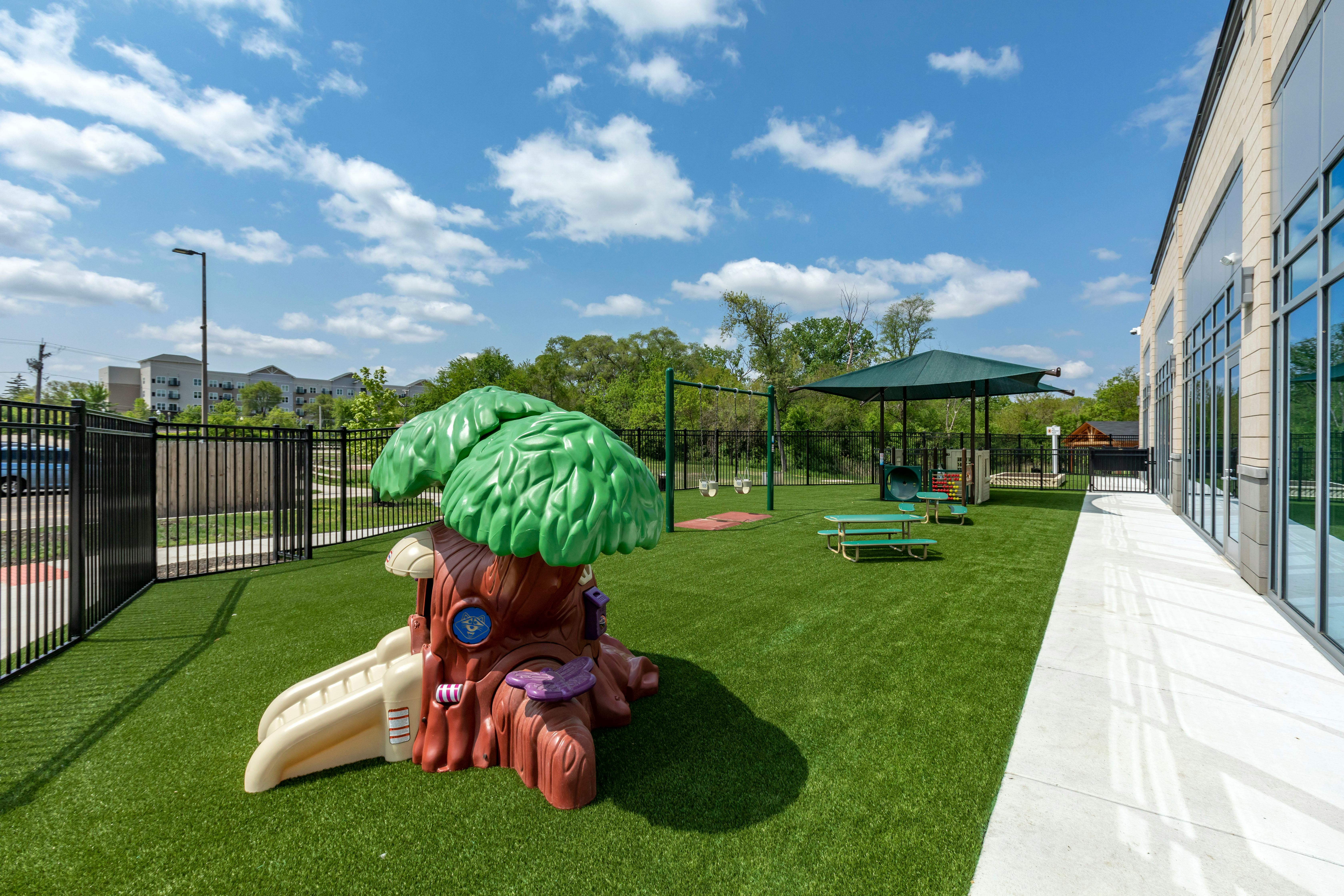 Outdoor playground with artificial turf, a tree-themed play structure, swings, and picnic tables