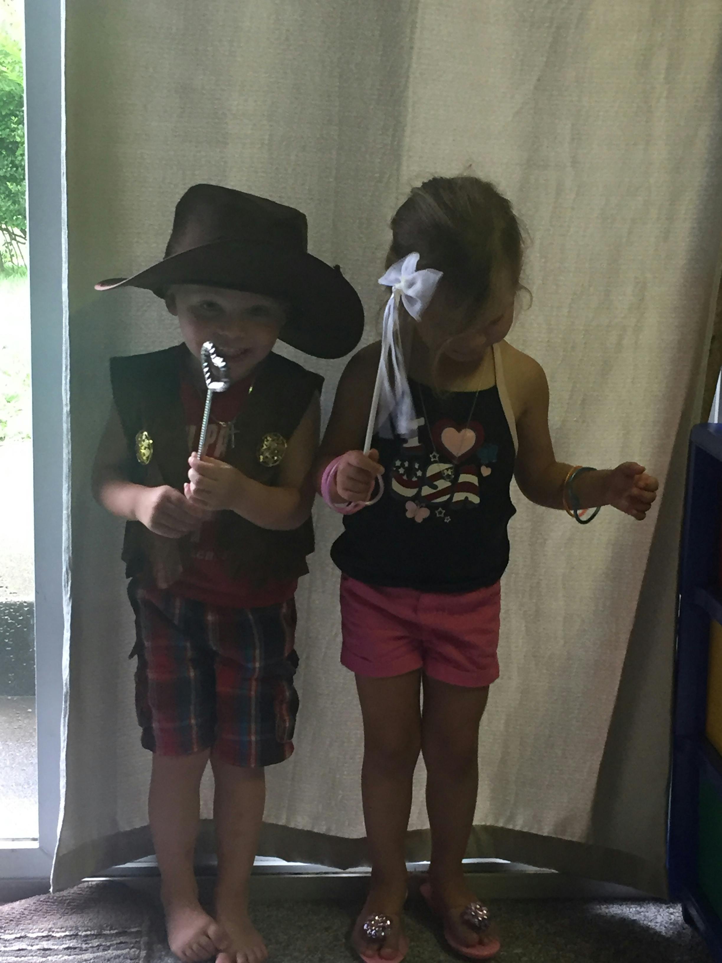 Child in cowboy hat and vest holding a toy, child in pink shorts holding a wand