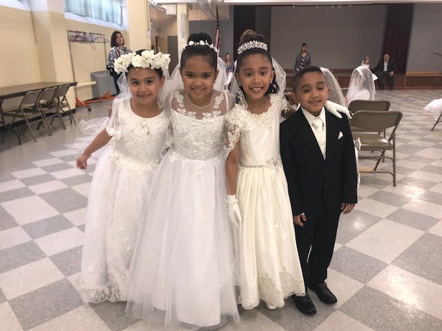 Three children in white dresses and one child in a suit standing together