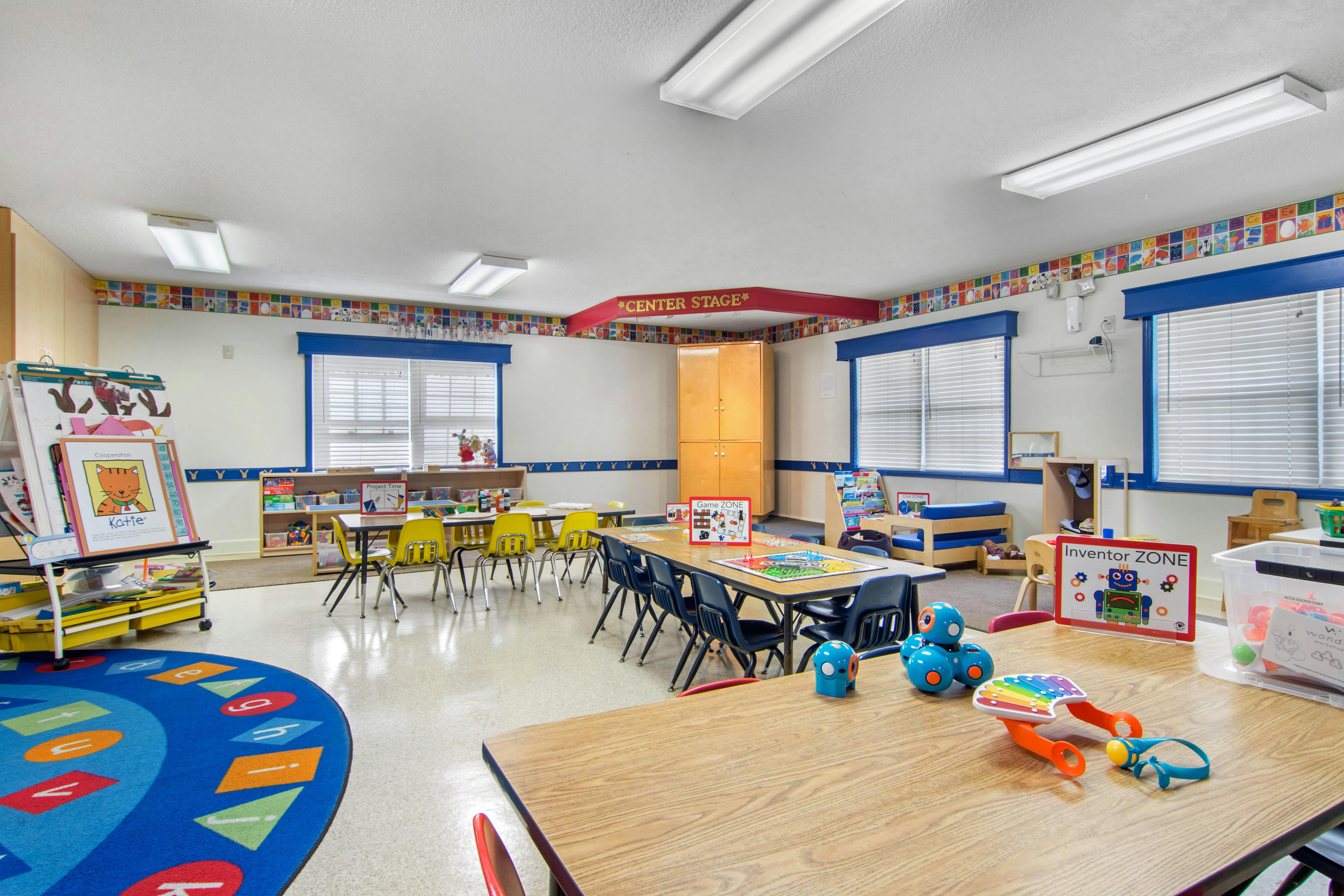 A classroom with tables and chairs, a play carpet, and various learning stations