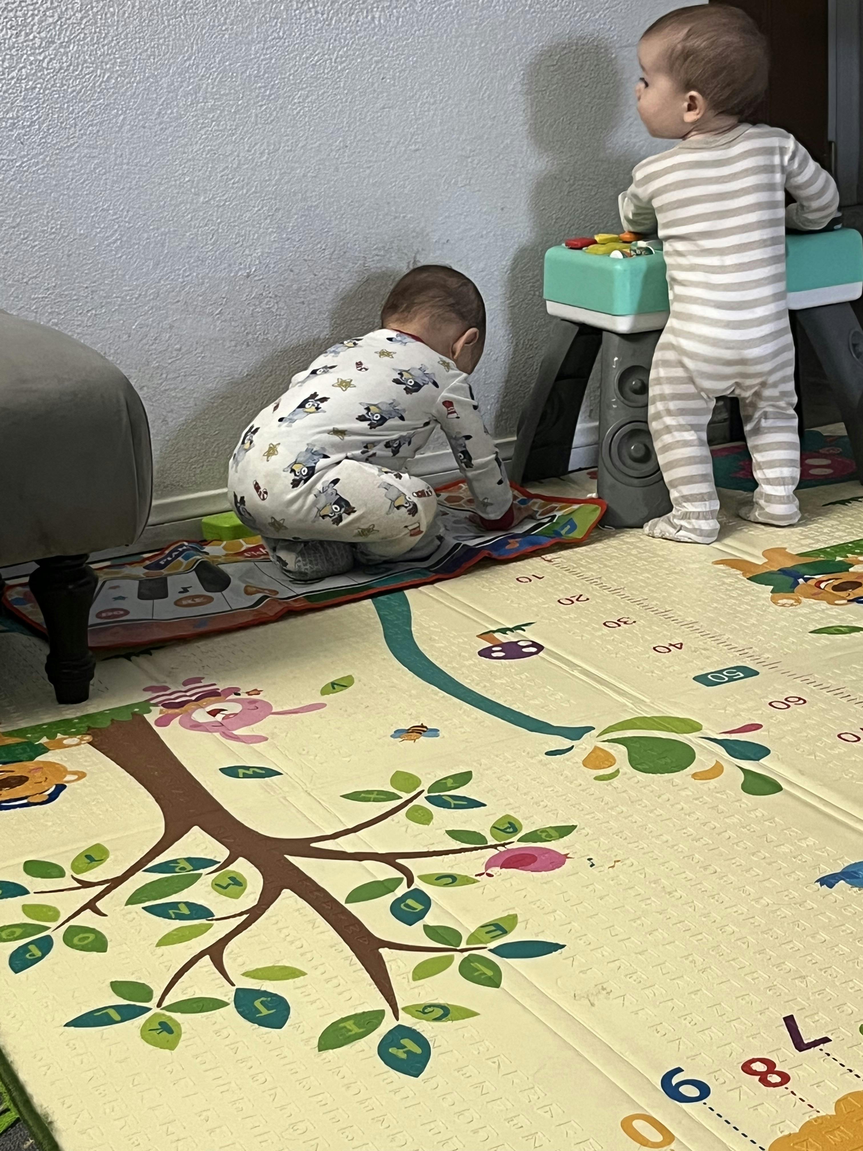 One child crawling on a playmat and another child standing at a toy