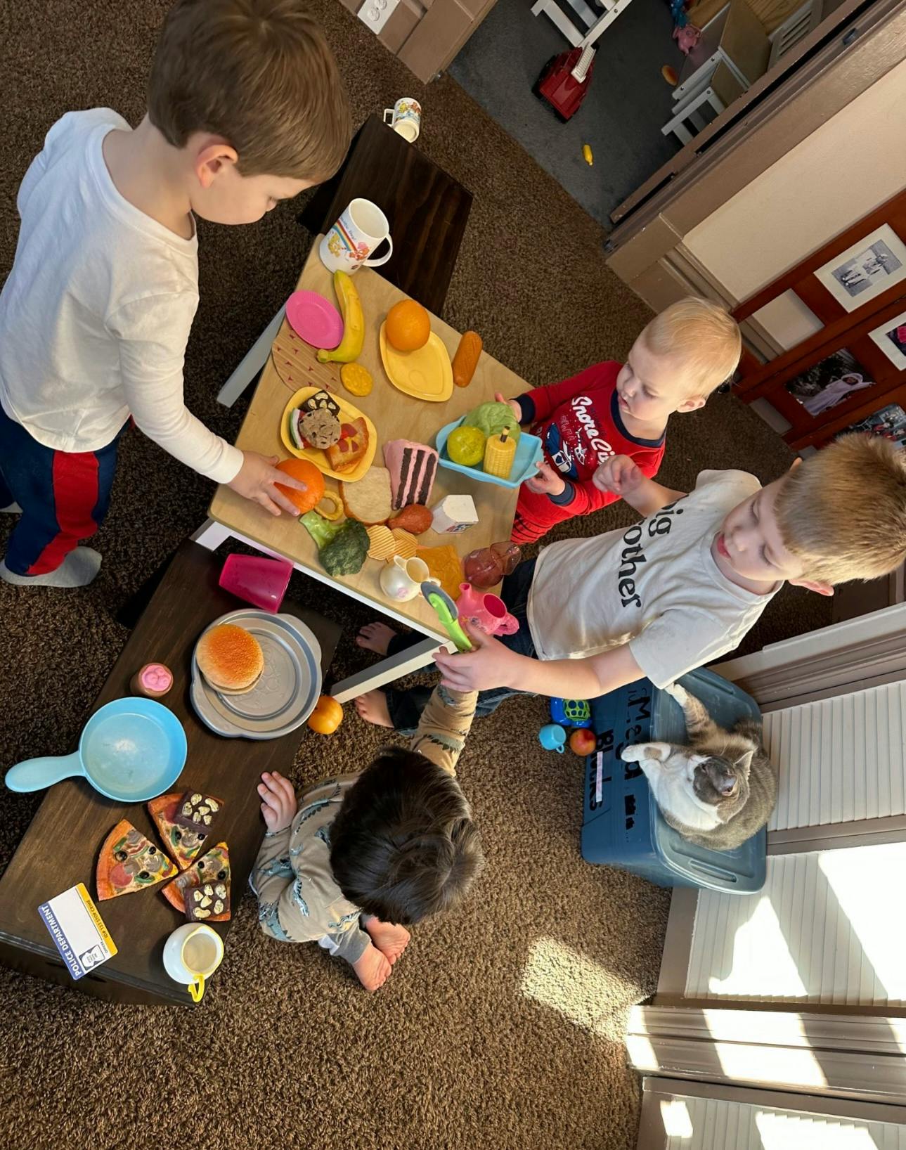 Three children playing with toy food on a table, one child on the floor