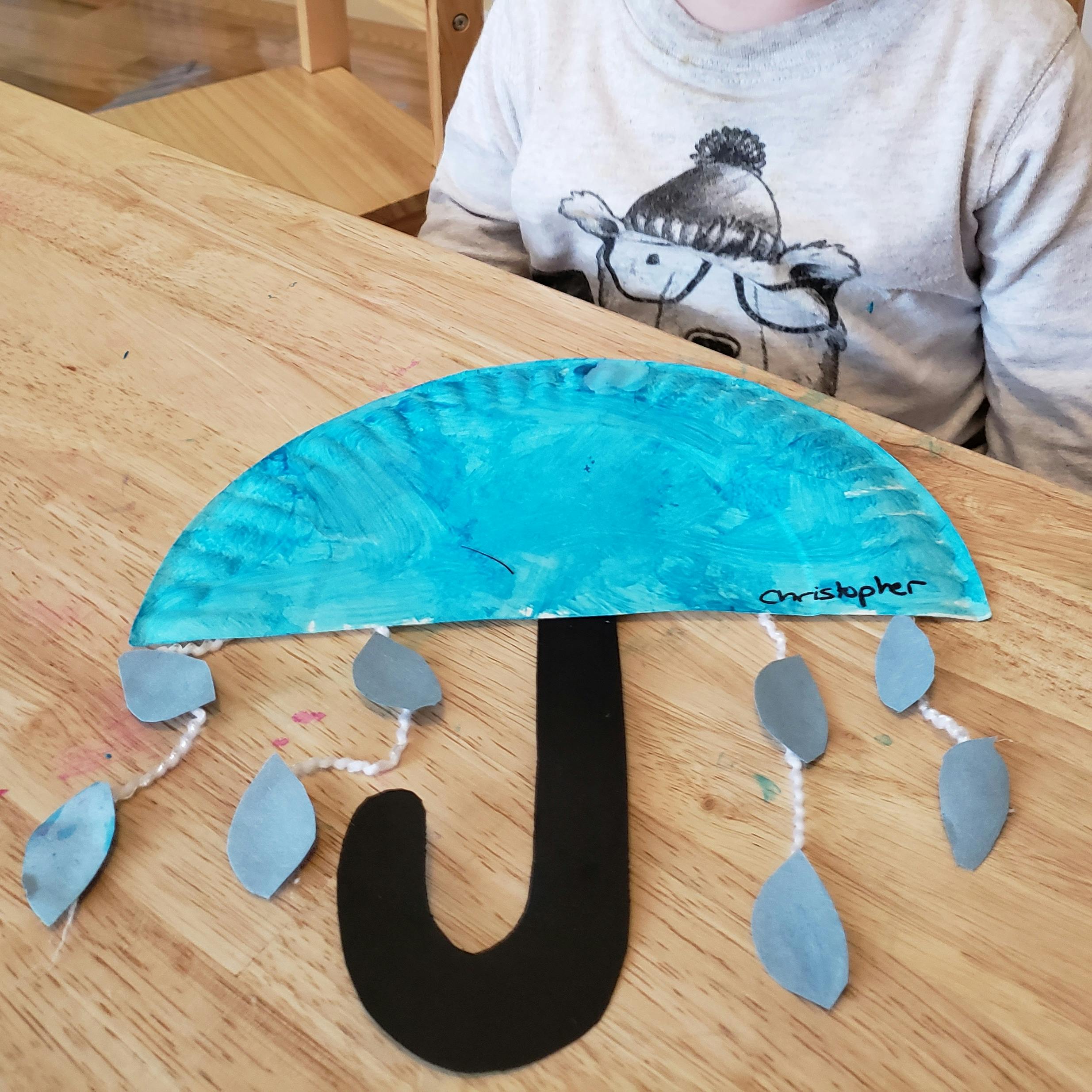 Child at a table with a blue paper plate umbrella craft with raindrops