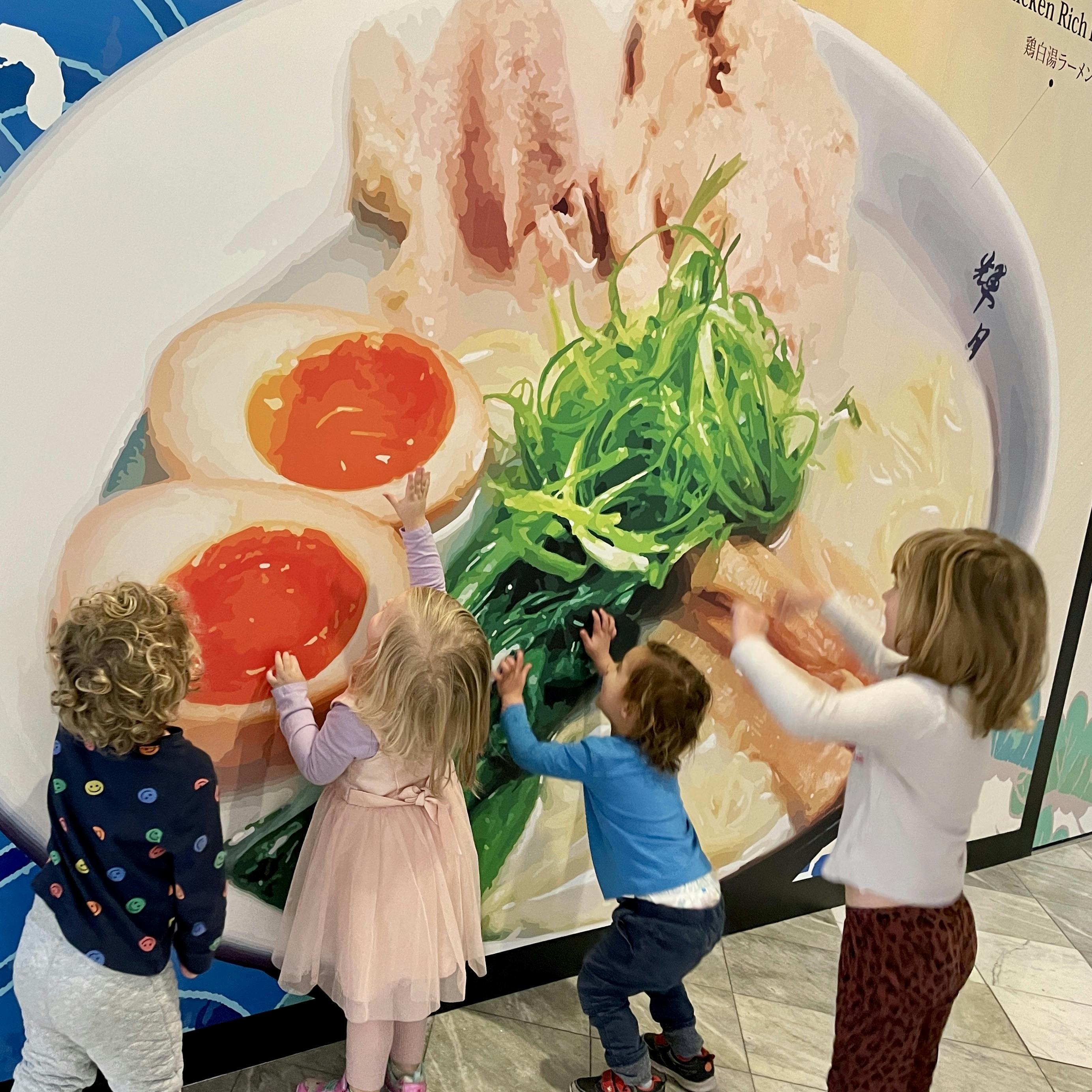 Four children touching a large wall mural of a bowl of ramen