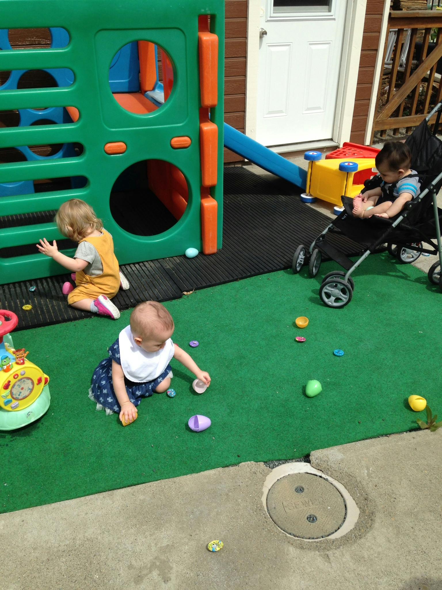 Three children playing on a green mat, one in a stroller
