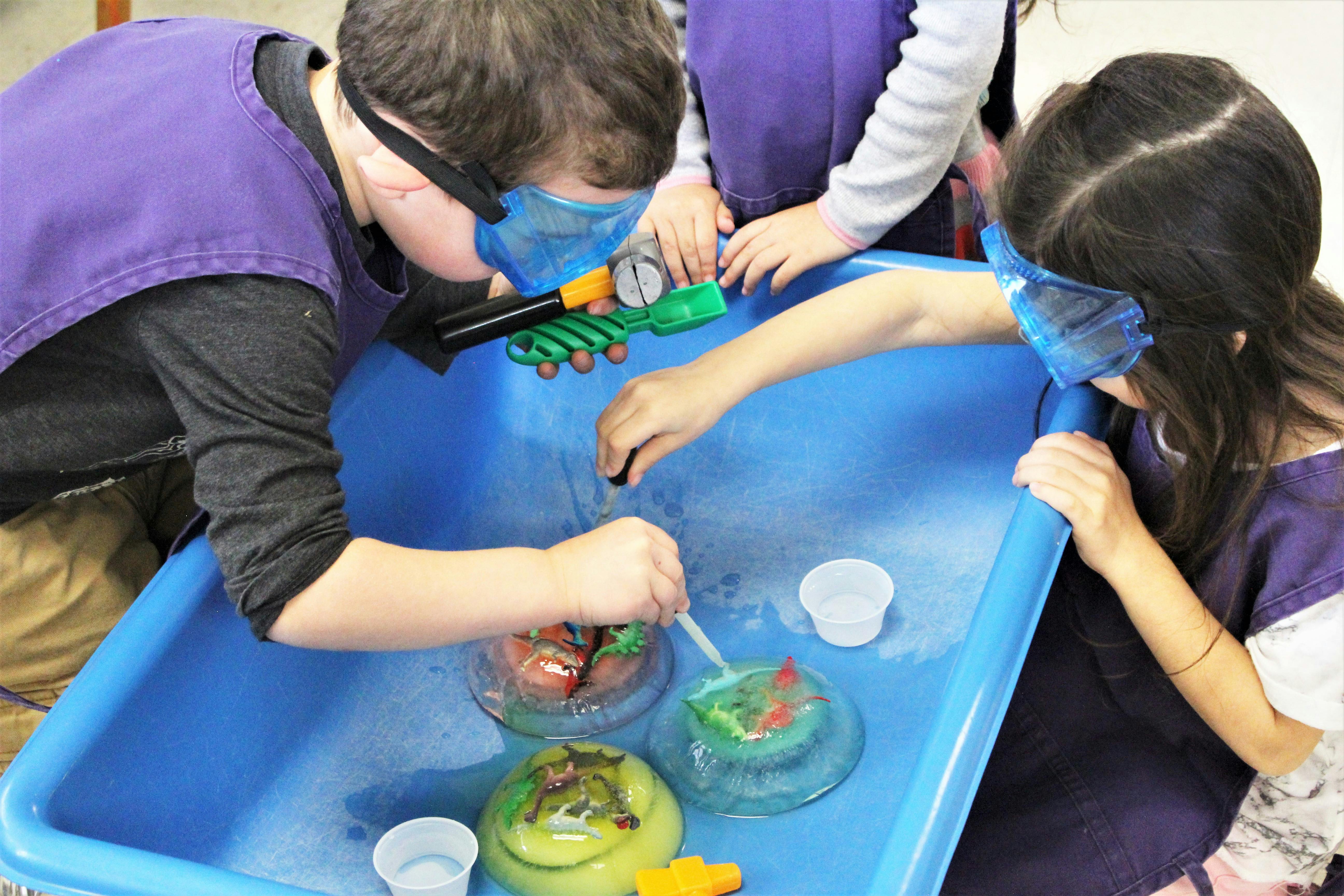 Children wearing safety goggles playing with ice and toy dinosaurs in a blue bin