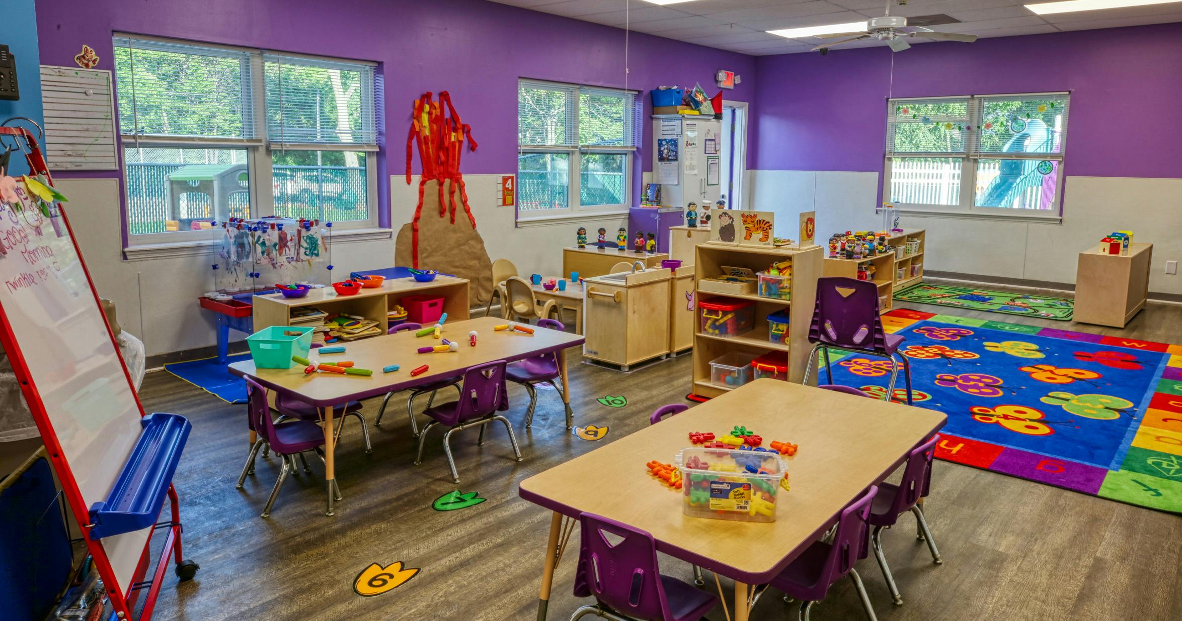 Colorful preschool classroom with purple walls, tables, chairs, and play areas