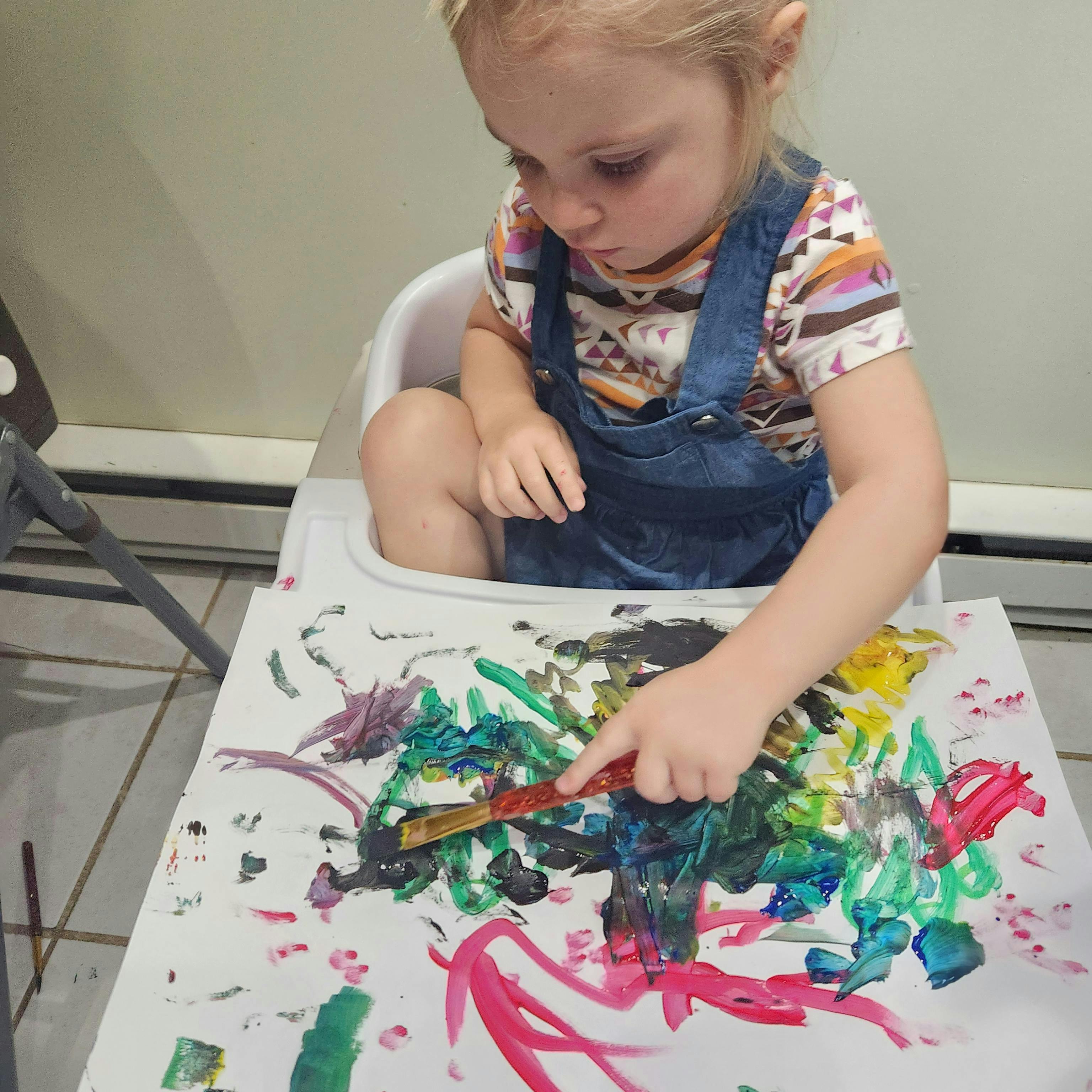 Child in a high chair painting on paper with a paintbrush