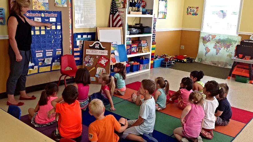 Person pointing at a chart with children sitting on a rug in a classroom