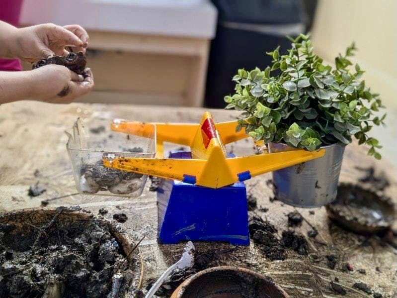Child's hands holding mud over a toy scale with a plant