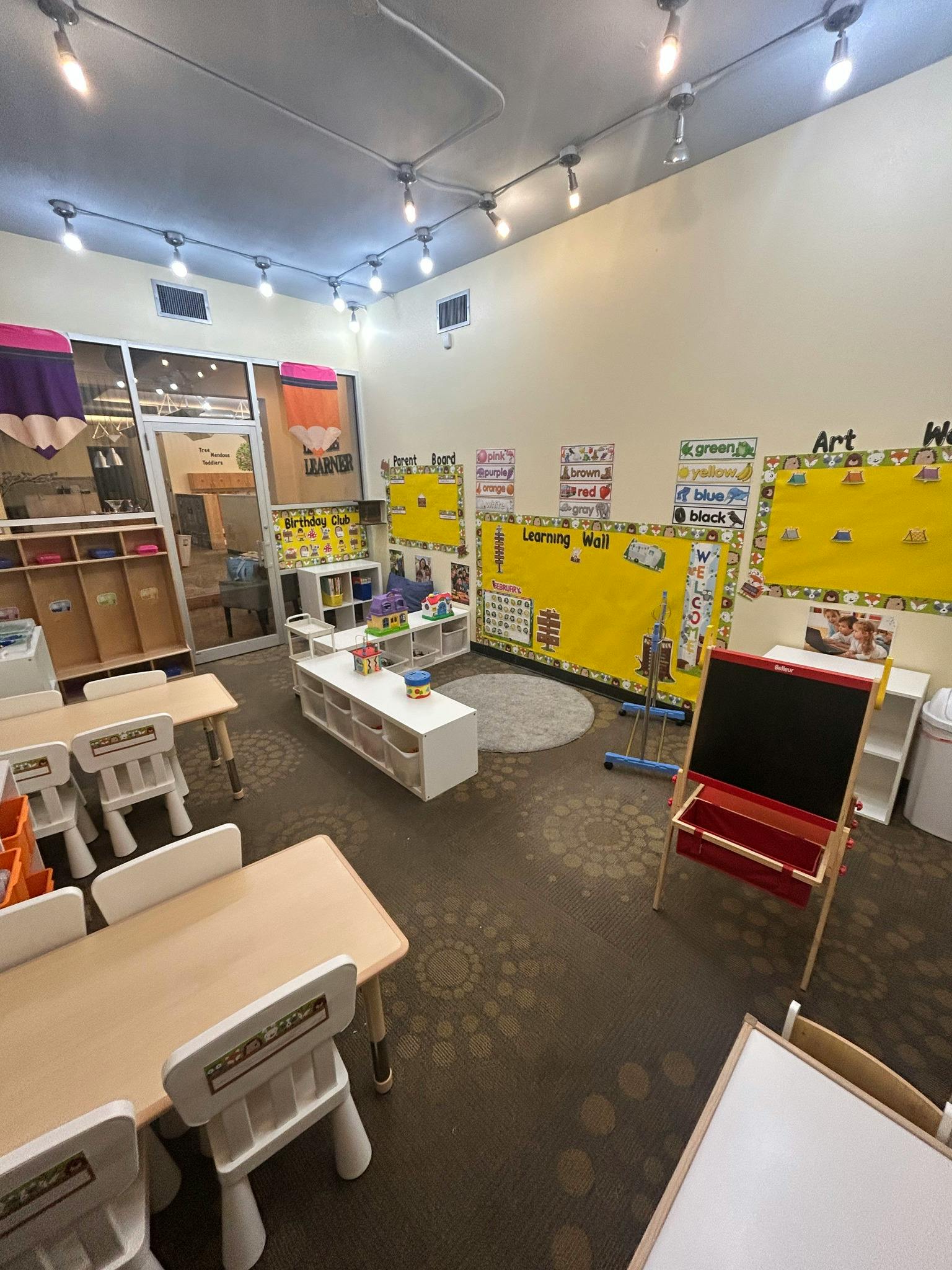 An empty classroom with small tables and chairs, a learning wall, and art supplies