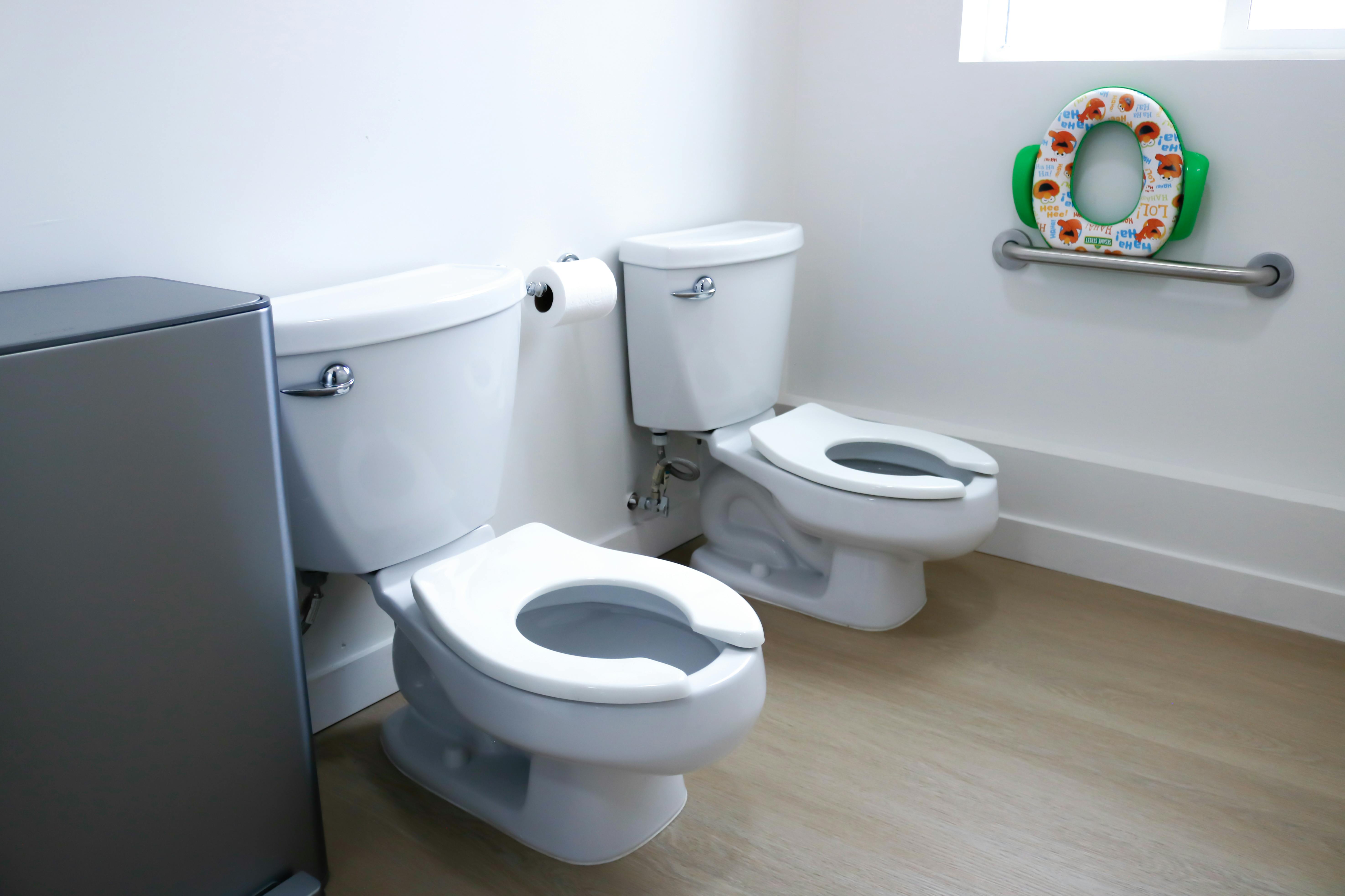 Two white toilets in a room, one with a child-sized seat hanging above it