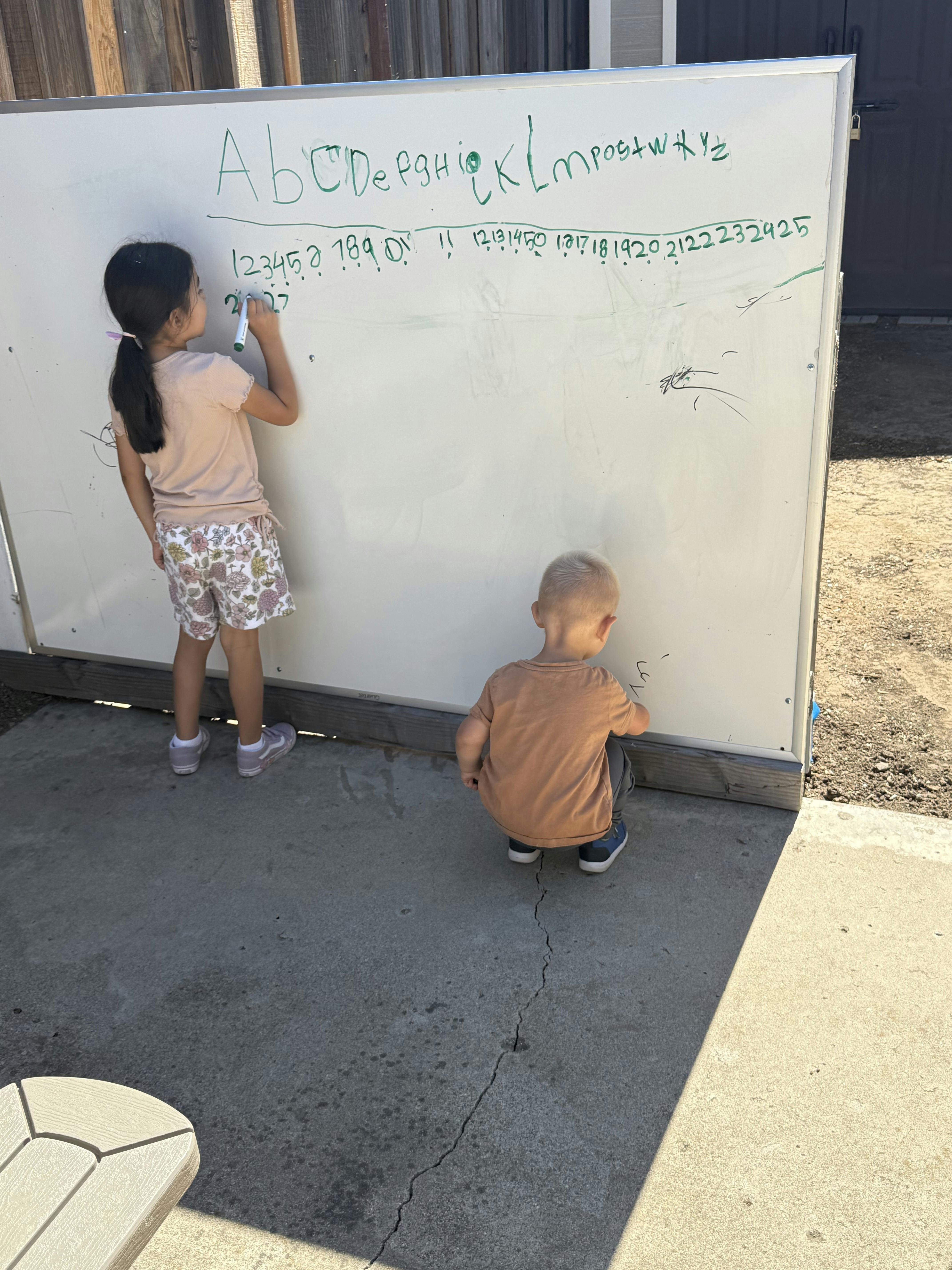 A child drawing on a large outdoor whiteboard with another child crouching nearby