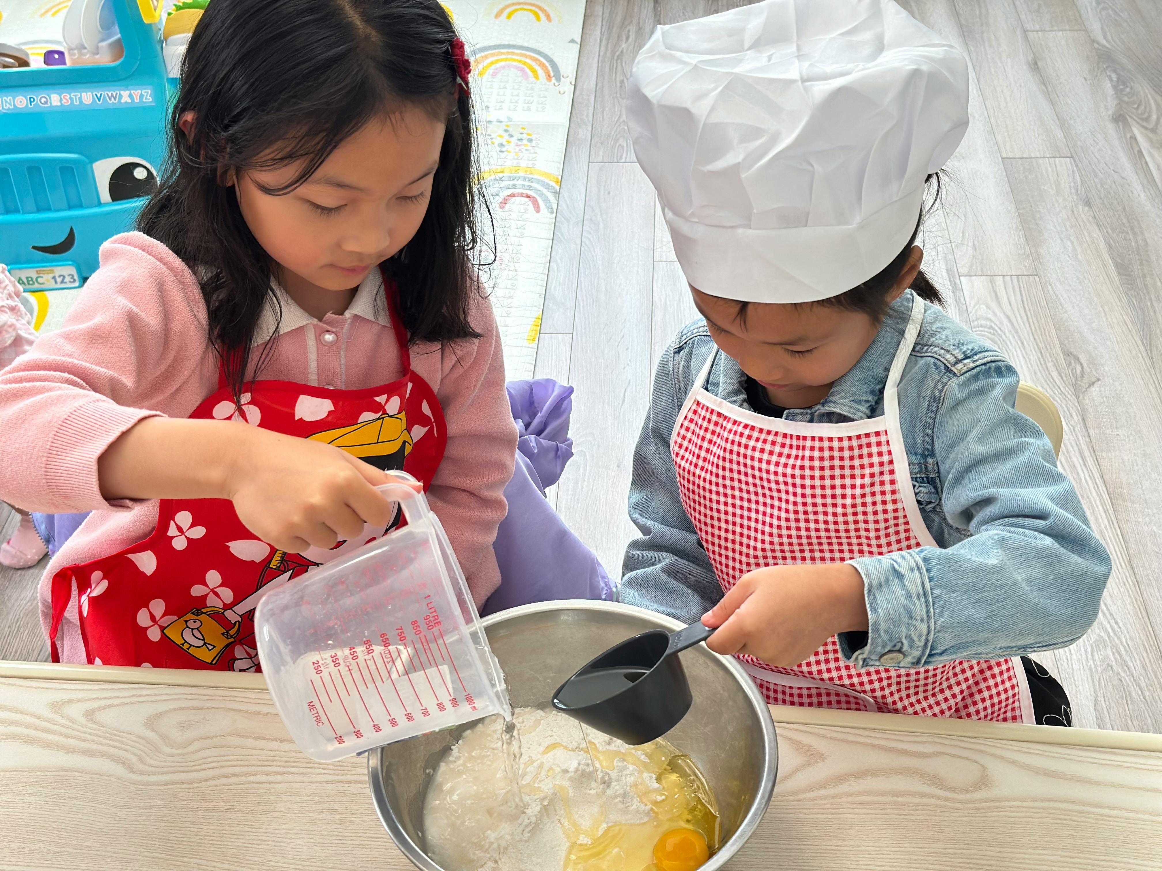 Two children wearing aprons pouring liquid into a mixing bowl of flour and eggs
