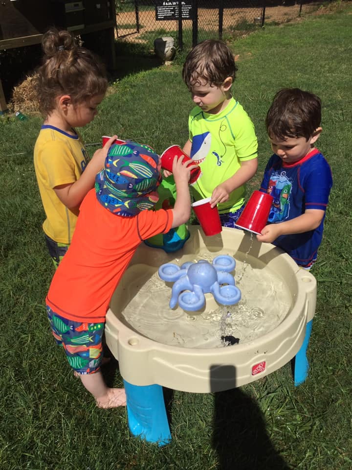 Three children playing with water and red cups at a water table