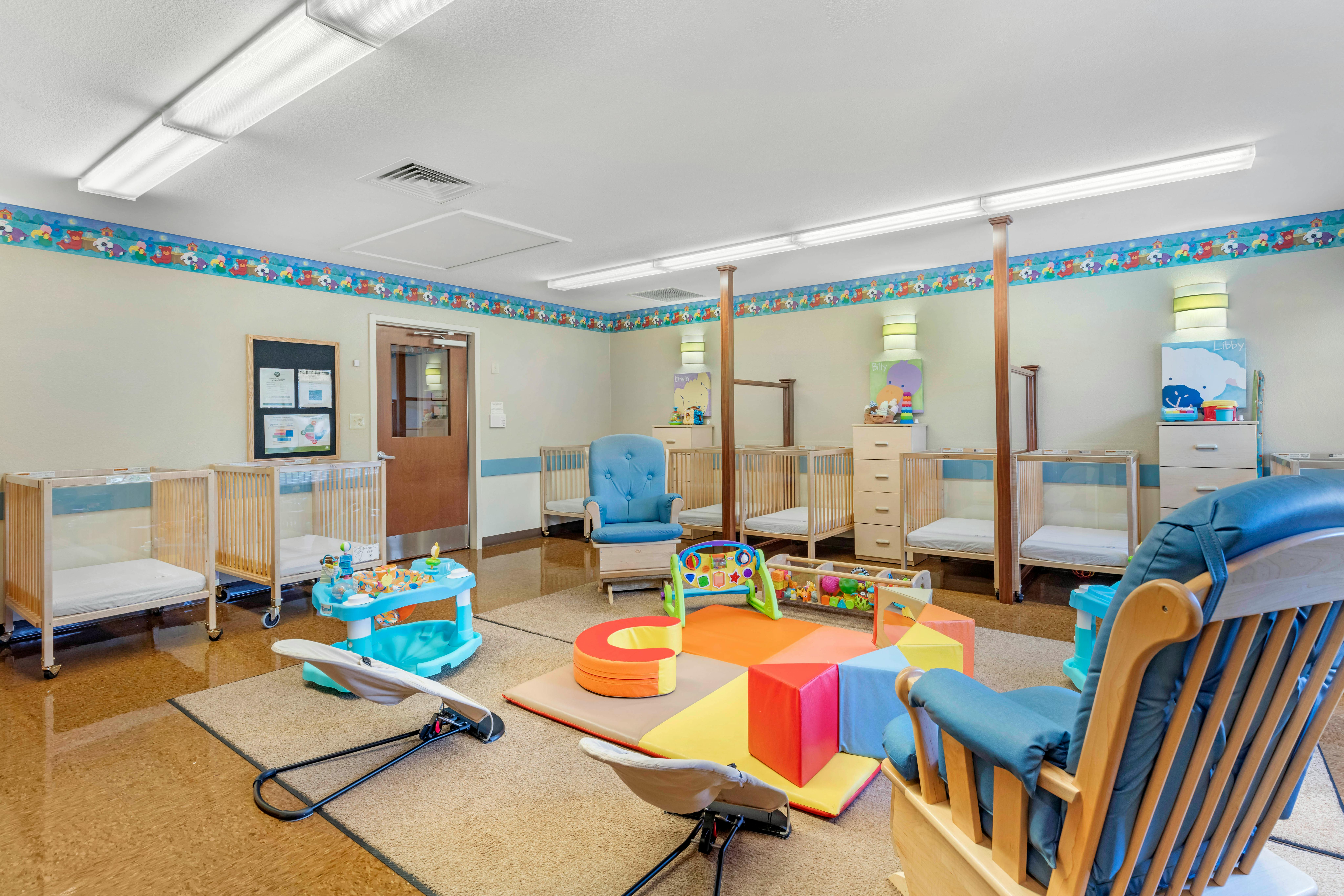 Baby room with multiple cribs, a blue rocking chair, and play mats with toys