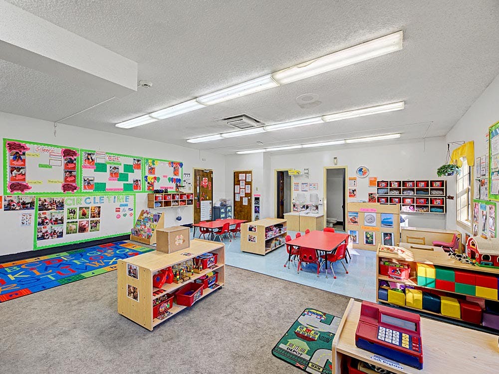 A classroom with small red tables and chairs, shelves with toys, and posters on the wall