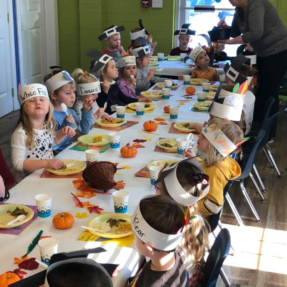 Children wearing paper hats sitting at a long table with plates of food