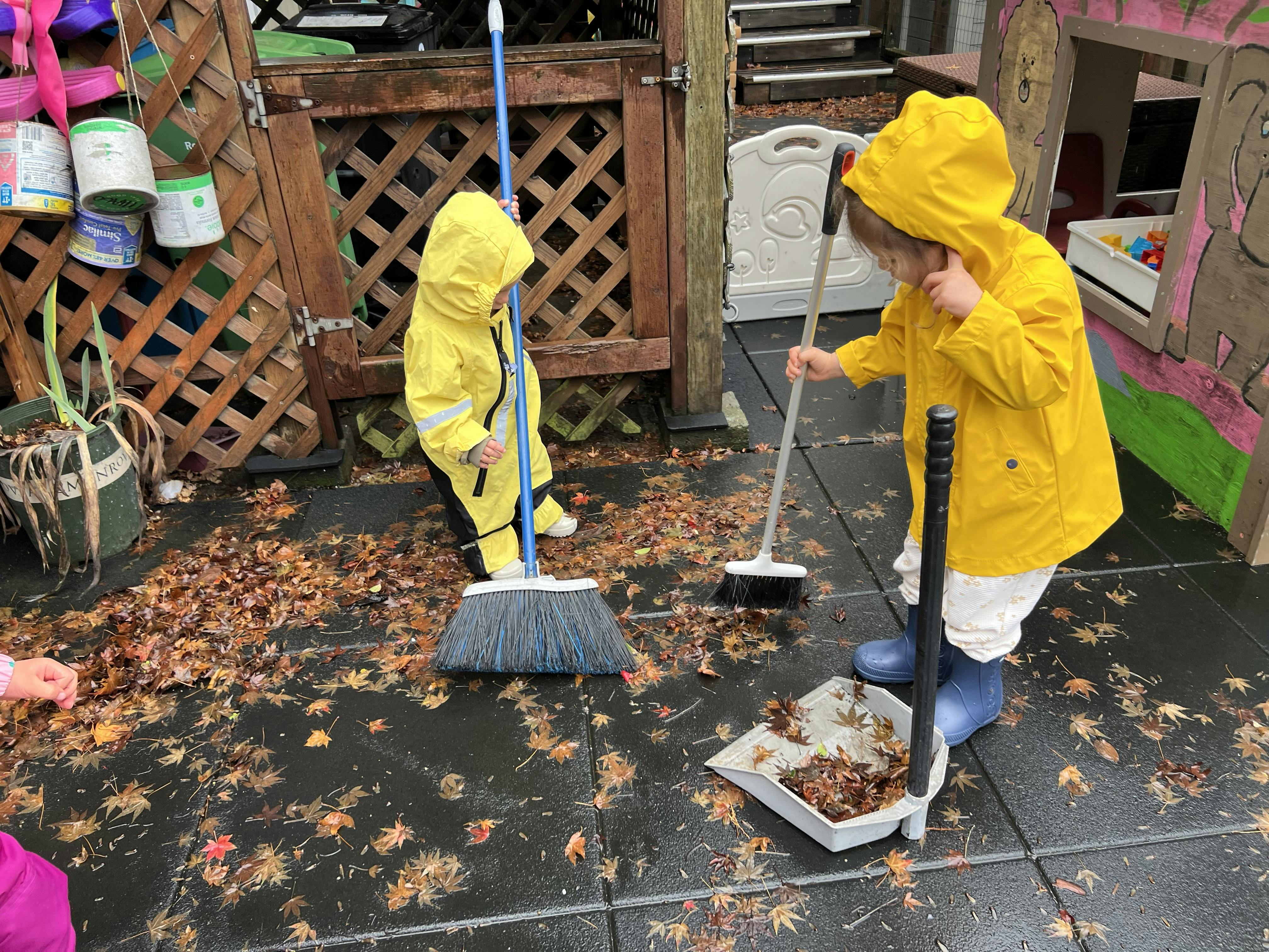 Two children in yellow raincoats sweeping leaves with brooms and a dustpan