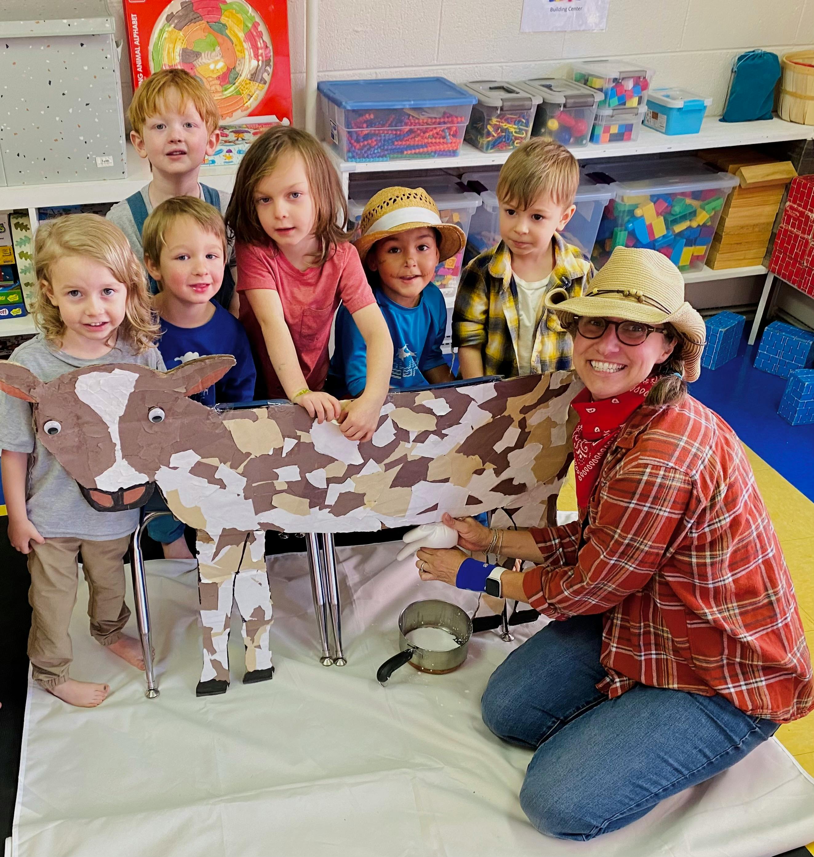 Adult and children gathered around a cow prop, adult milking the cow