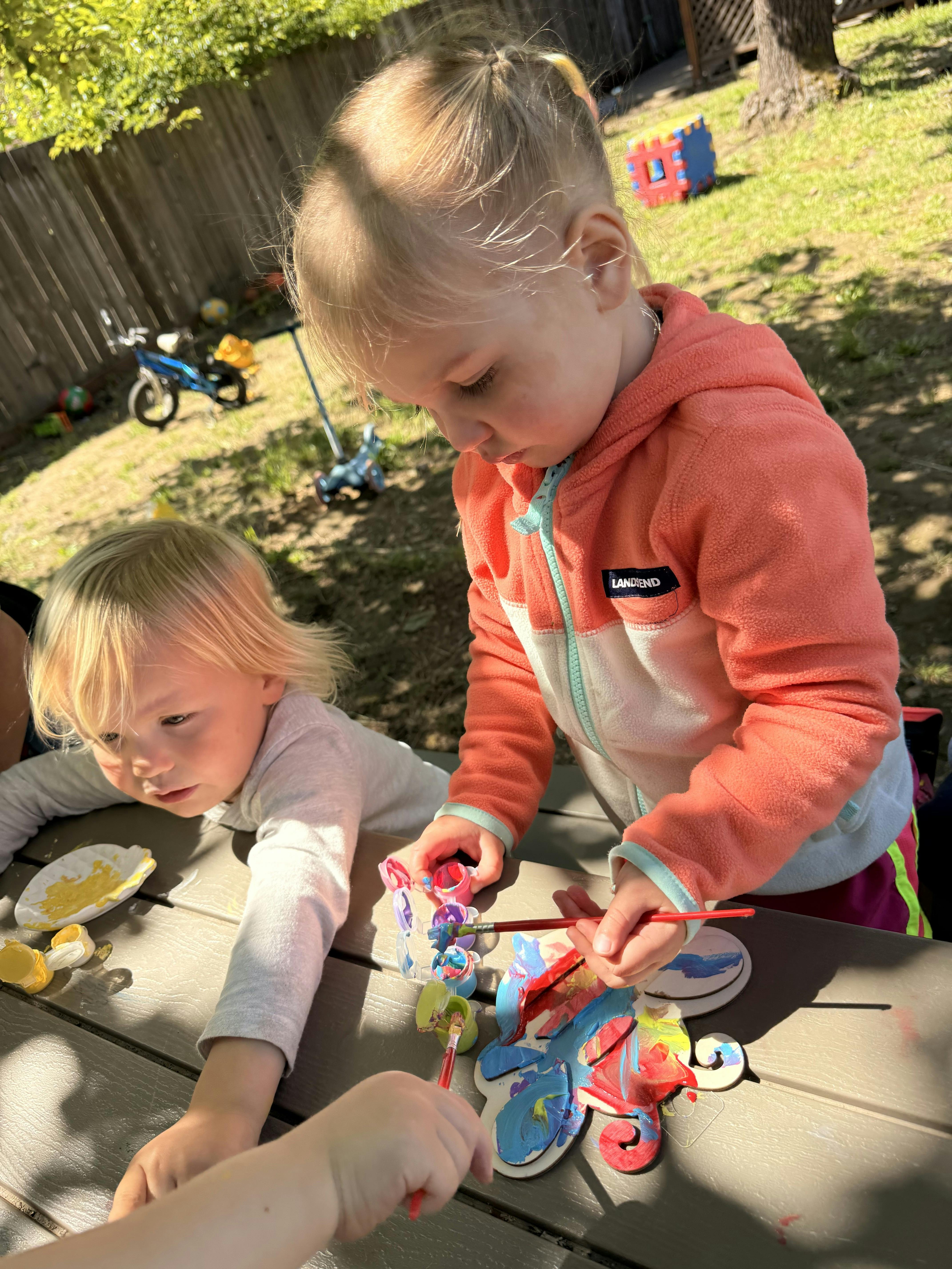 Two children painting wooden cutouts at an outdoor table