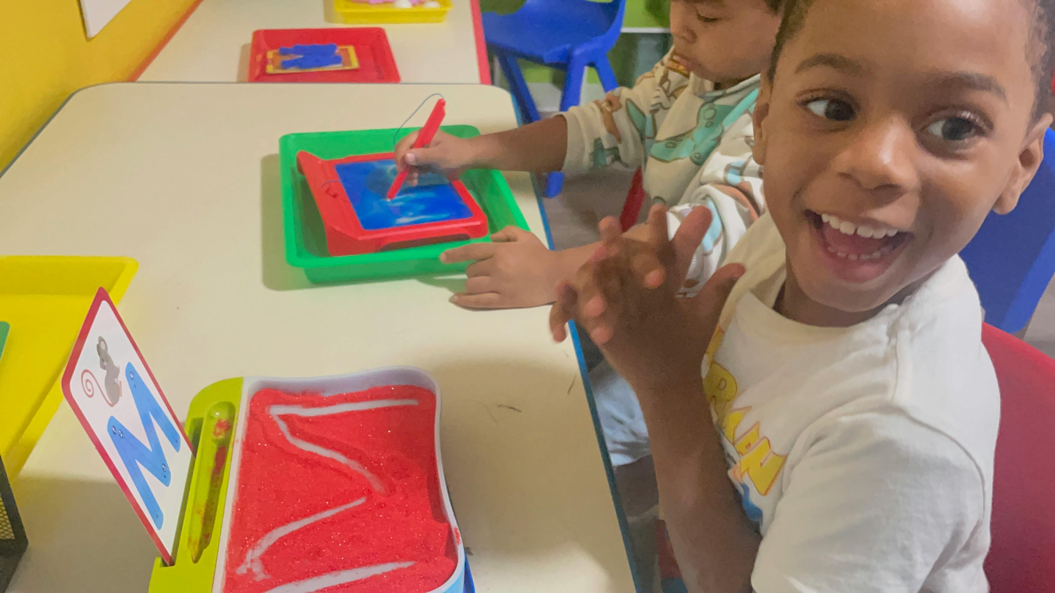 Child smiling at the camera, another child drawing on a magnetic board