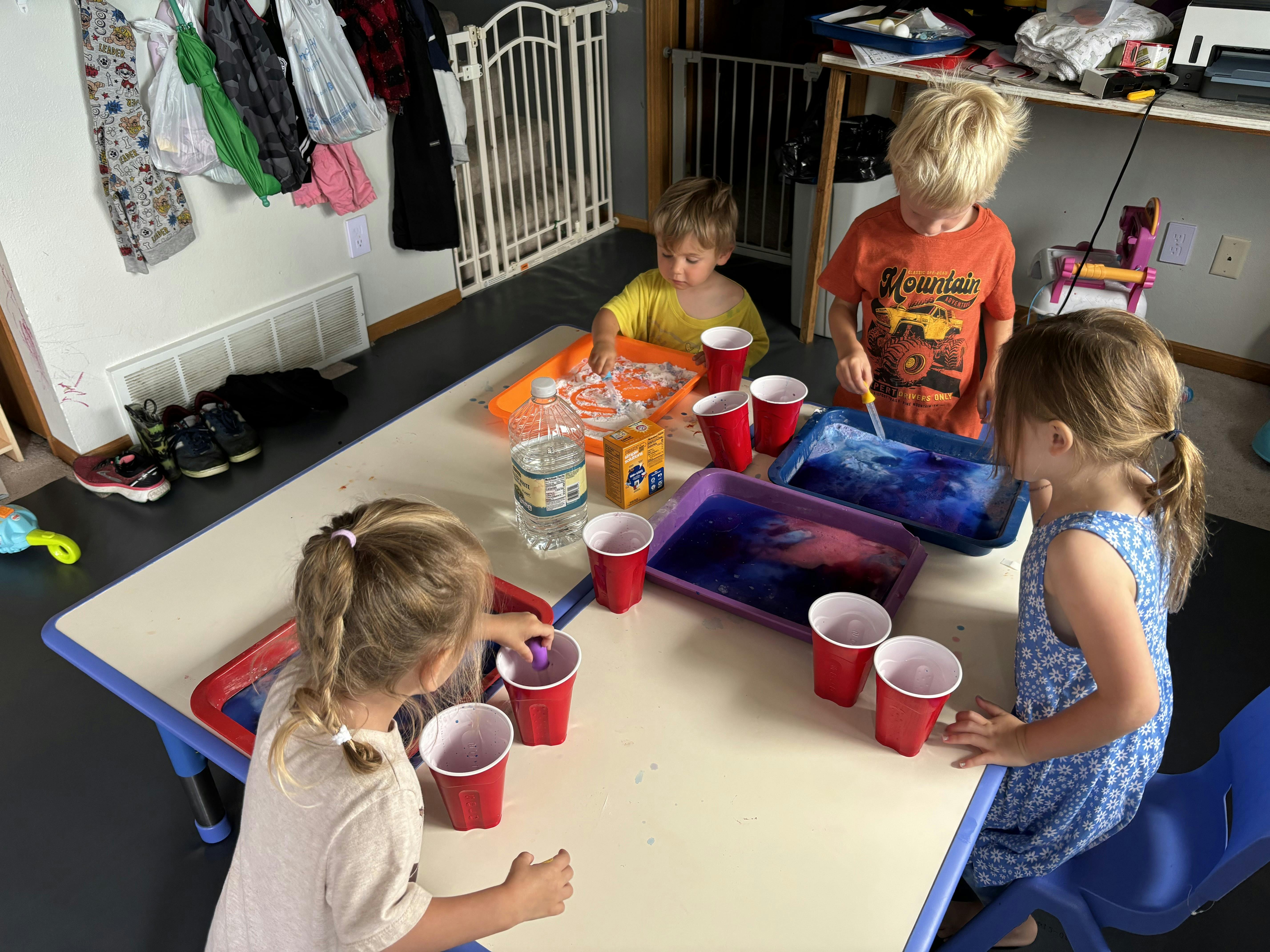 Four children at a table with red cups and trays of colored liquid