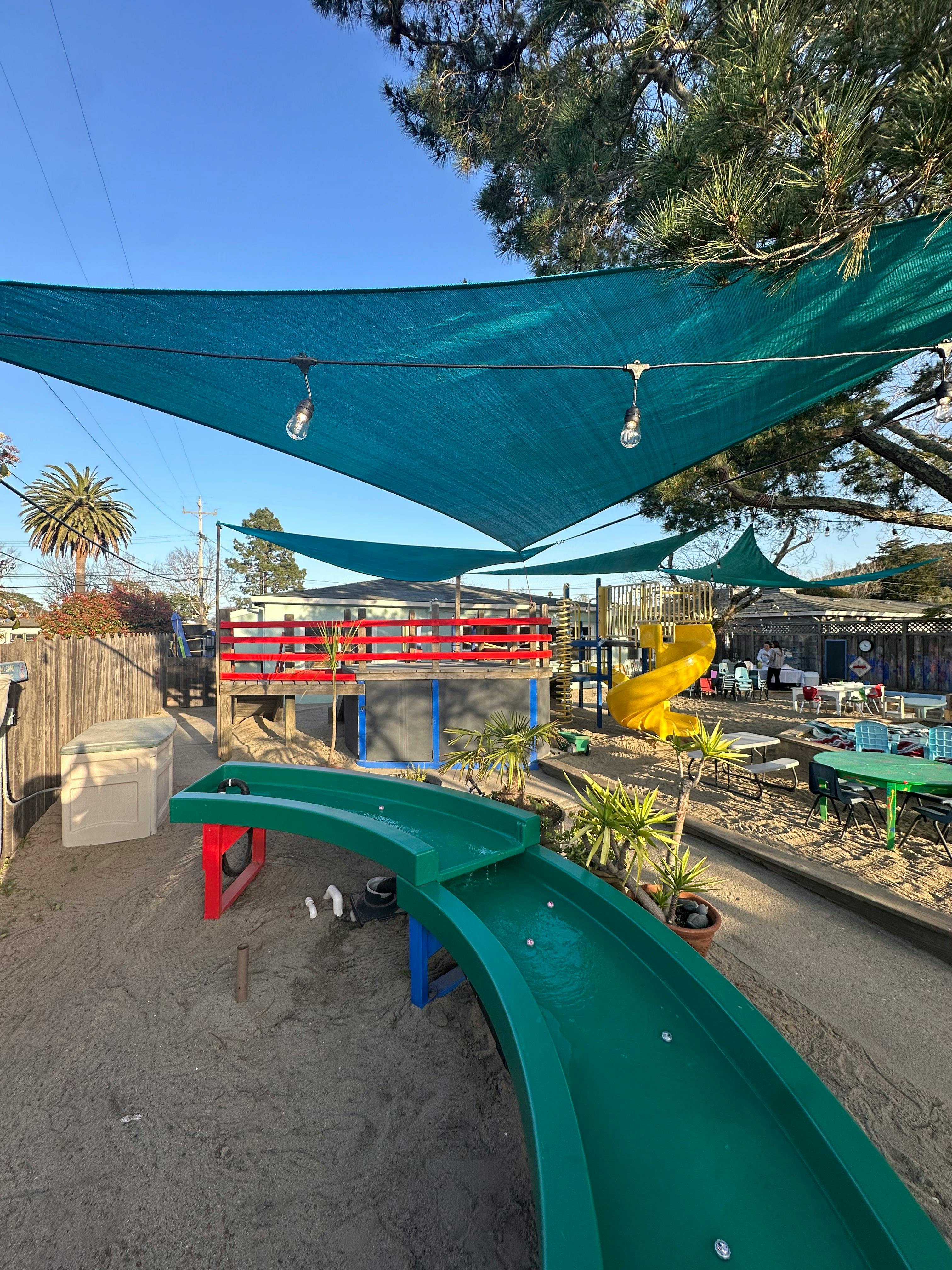 Outdoor play area with green shade sails, a yellow slide, and a green water table