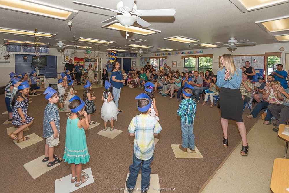 Adults watching children in blue hats standing on mats in a room