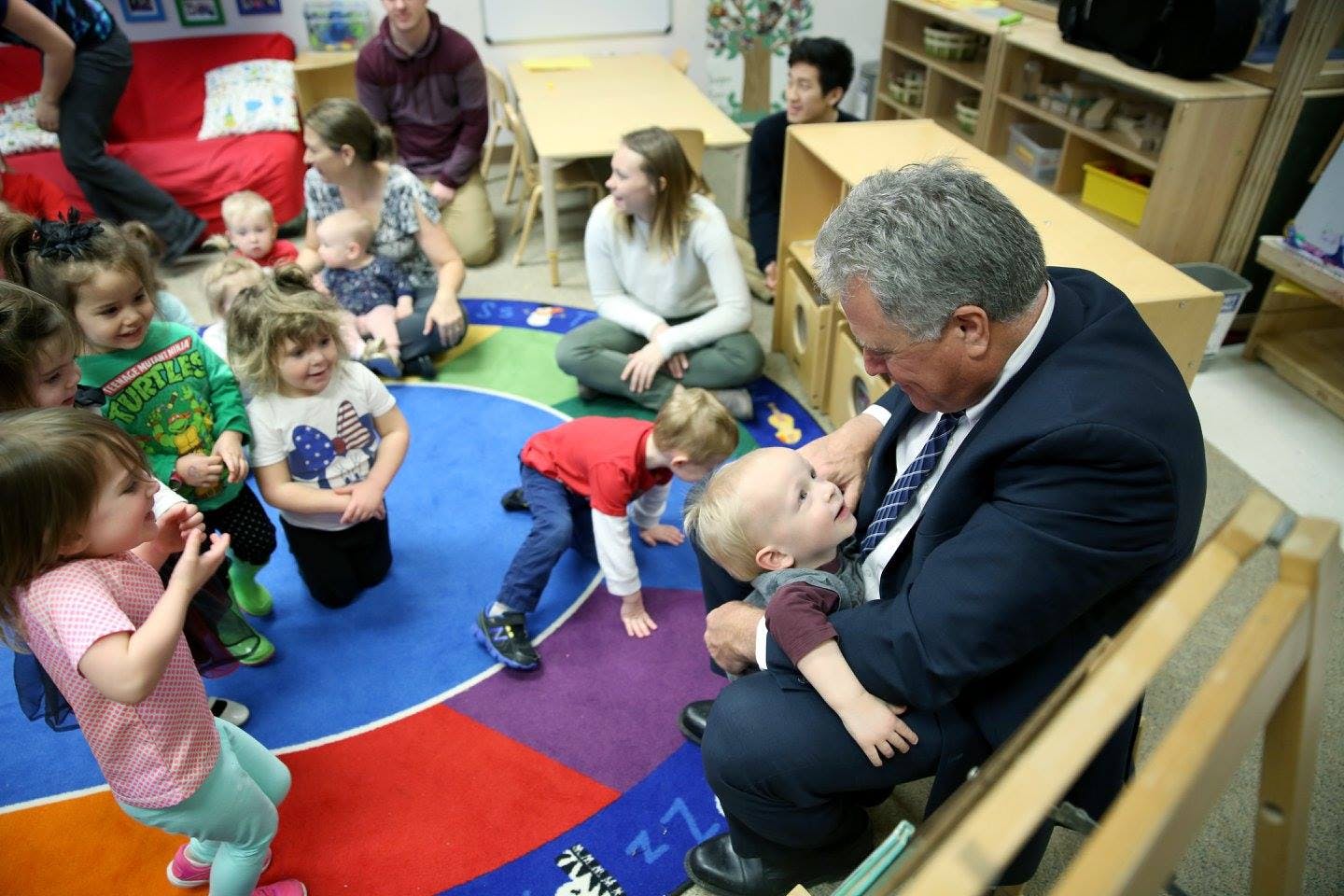 Adult holding a child in a classroom with other adults and children