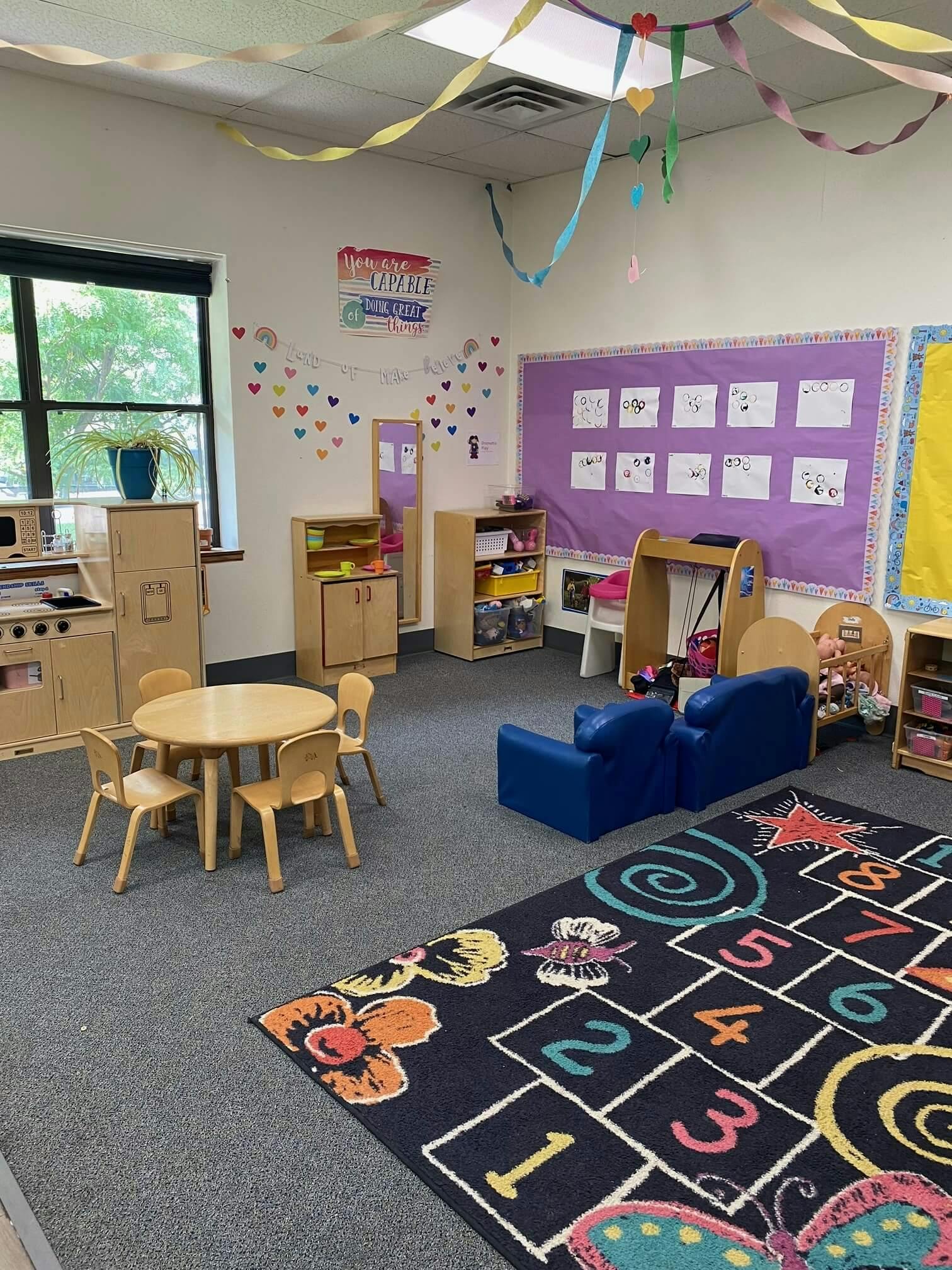 Children's classroom with small table and chairs, play kitchen, and hopscotch rug
