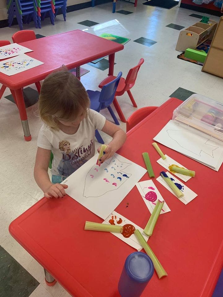 Child painting with celery stalks at a red table in a classroom